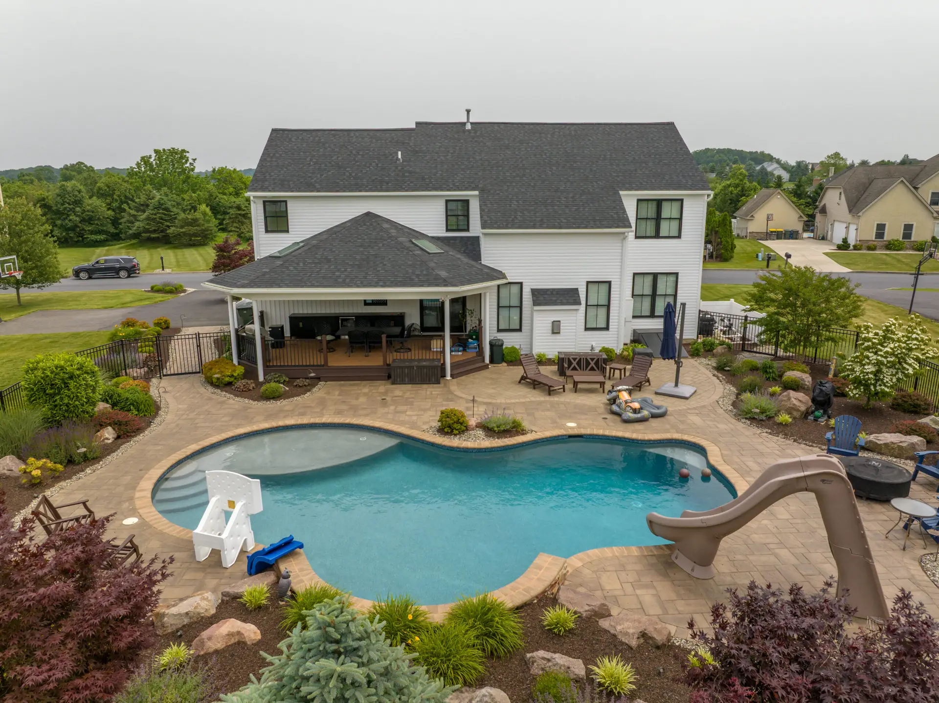 Backyard view of a large beige house with a curved pool, lounge chairs, and a waterfall slide, surrounded by landscaped plants and a paved patio.
