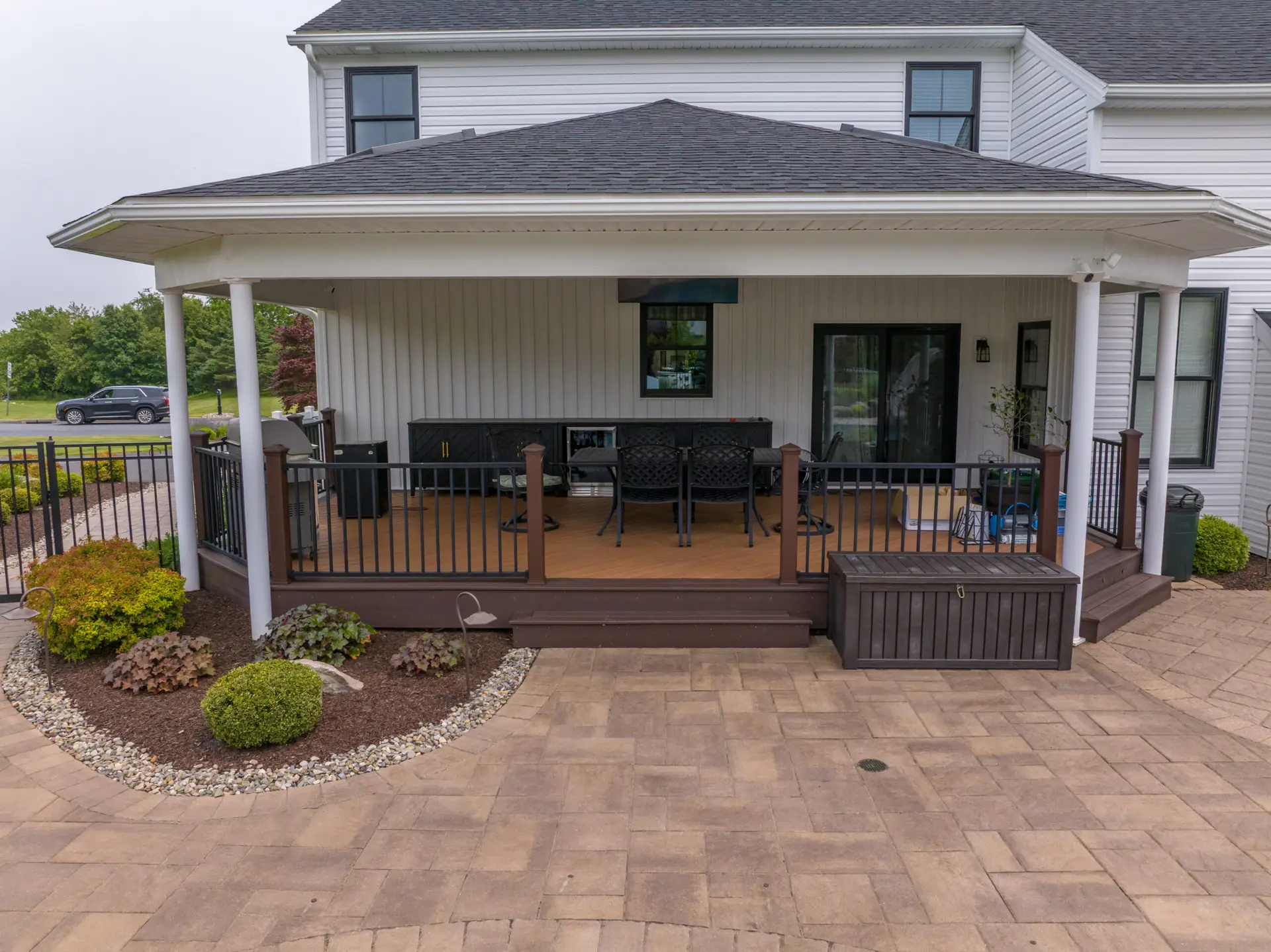 Backyard patio under a covered porch with dark wicker furniture, grill, and potted plants beside a white house.