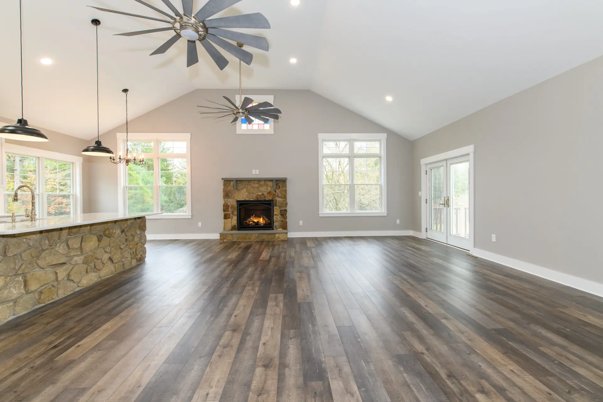 Open living space with vaulted white ceiling, stone fireplace, large windows, and dark wood floors; kitchen island on left and French doors on right.