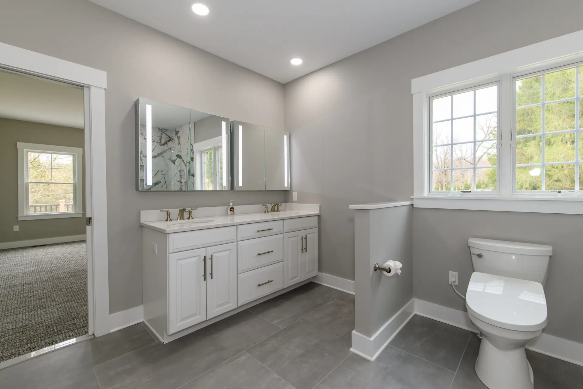 Modern bathroom with a white double-sink vanity, mirrored cabinets, and gray walls under recessed lighting facing a large window Fall view outside.