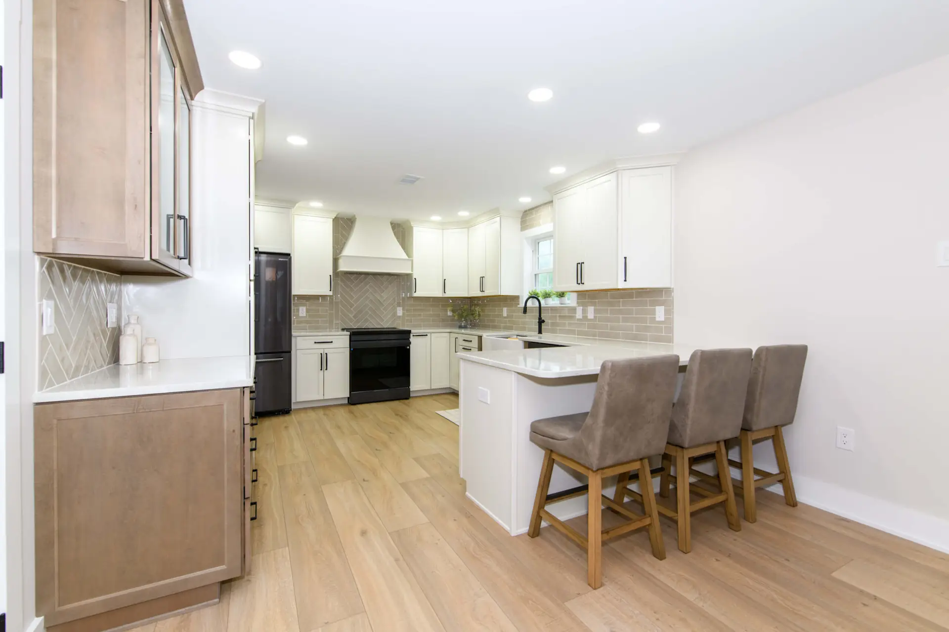 Modern kitchen with white cabinets, light tile backsplash, and a centered island with three beige stools; hardwood floors and stainless steel appliances visible.