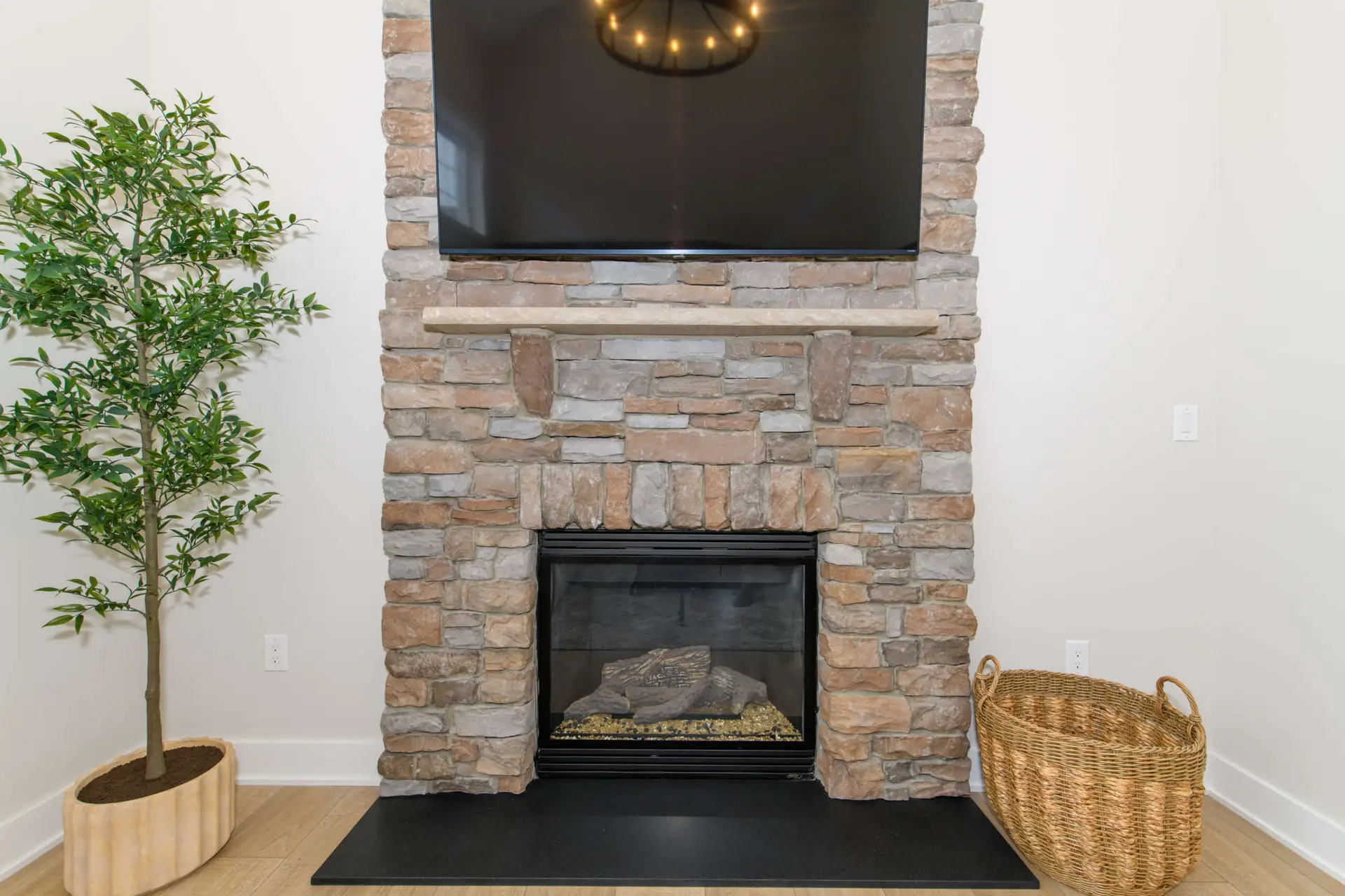 Stone fireplace with a mounted flat-screen TV above and a wooden mantel, flanked by a green plant on the left and a wicker basket on the right in a living room.