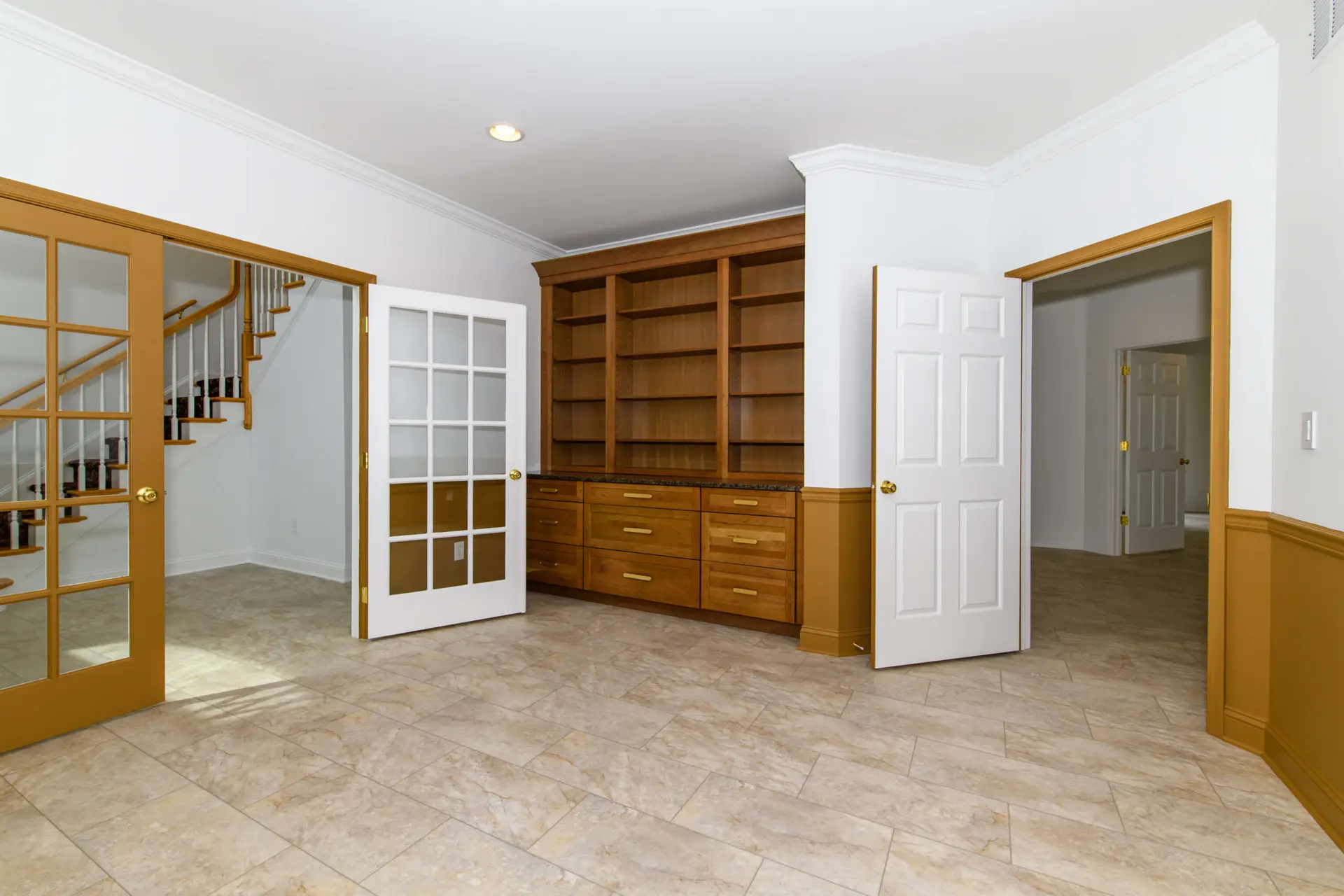 Interior view of a room featuring built-in wooden shelves and drawers, white doors, and a glass-panel French door on the left, with a staircase visible through the door.