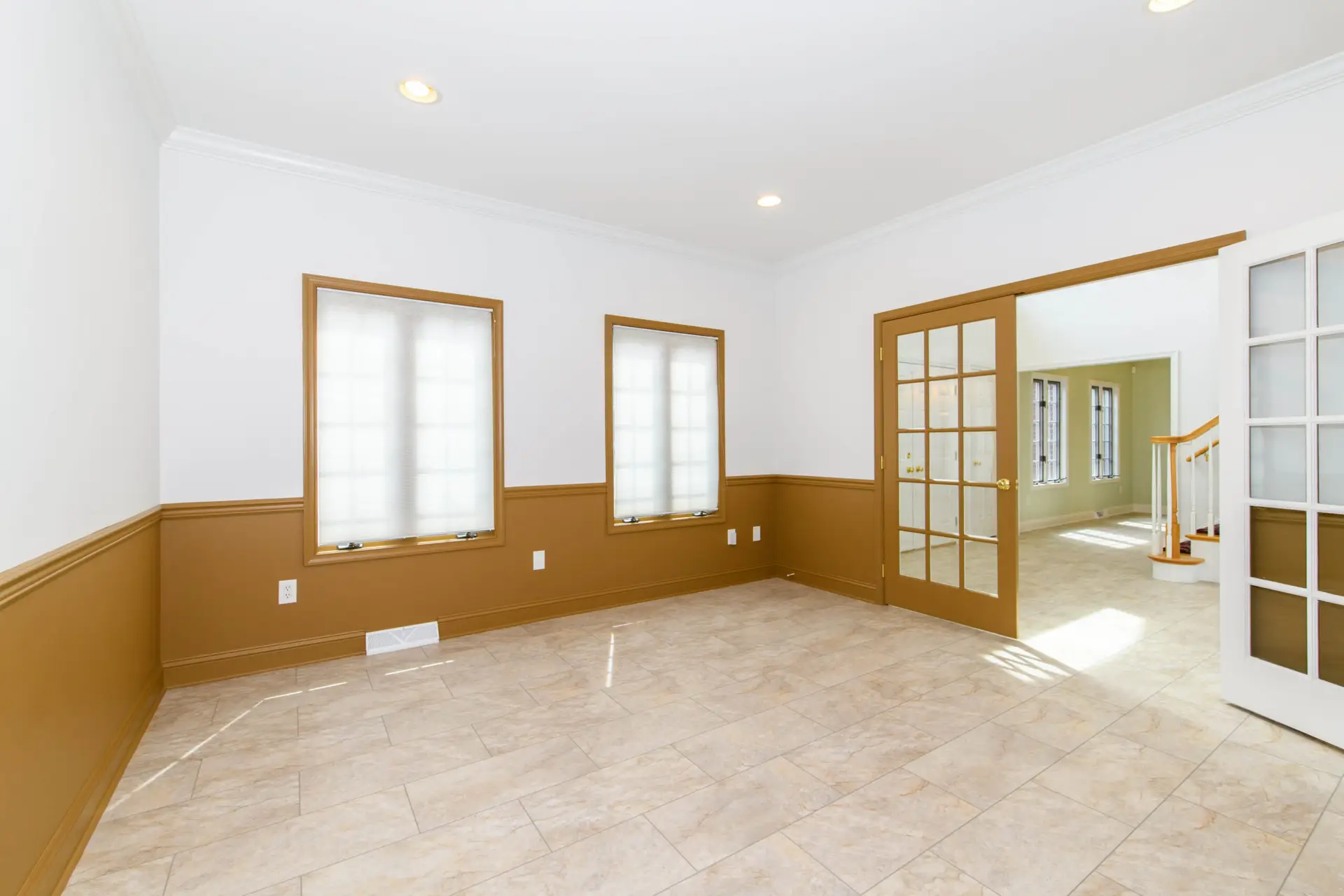 Empty sunlit interior room with beige tile floor, white walls, and brown wainscoting; glass French doors lead to a connected space with a staircase visible in the background.