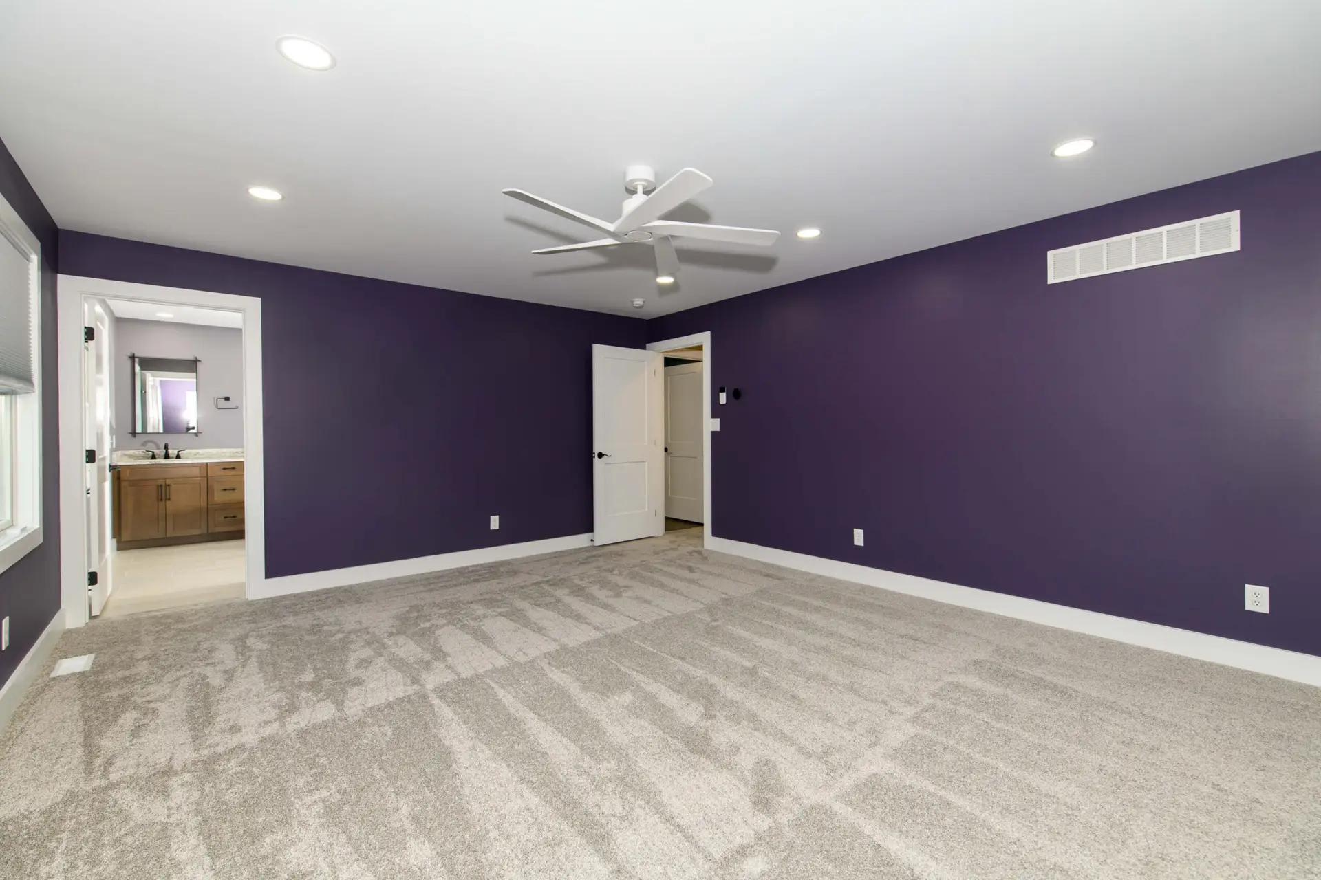 Empty purple-walled bedroom with gray carpet, white baseboards, and a white ceiling fan; doorway to a bathroom visible on the left and a closed door on the back wall.