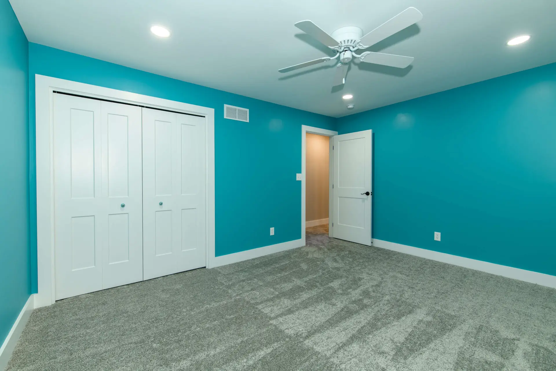 Turquoise room with white double closet doors, open entry door, ceiling fan, and gray carpeted floor.