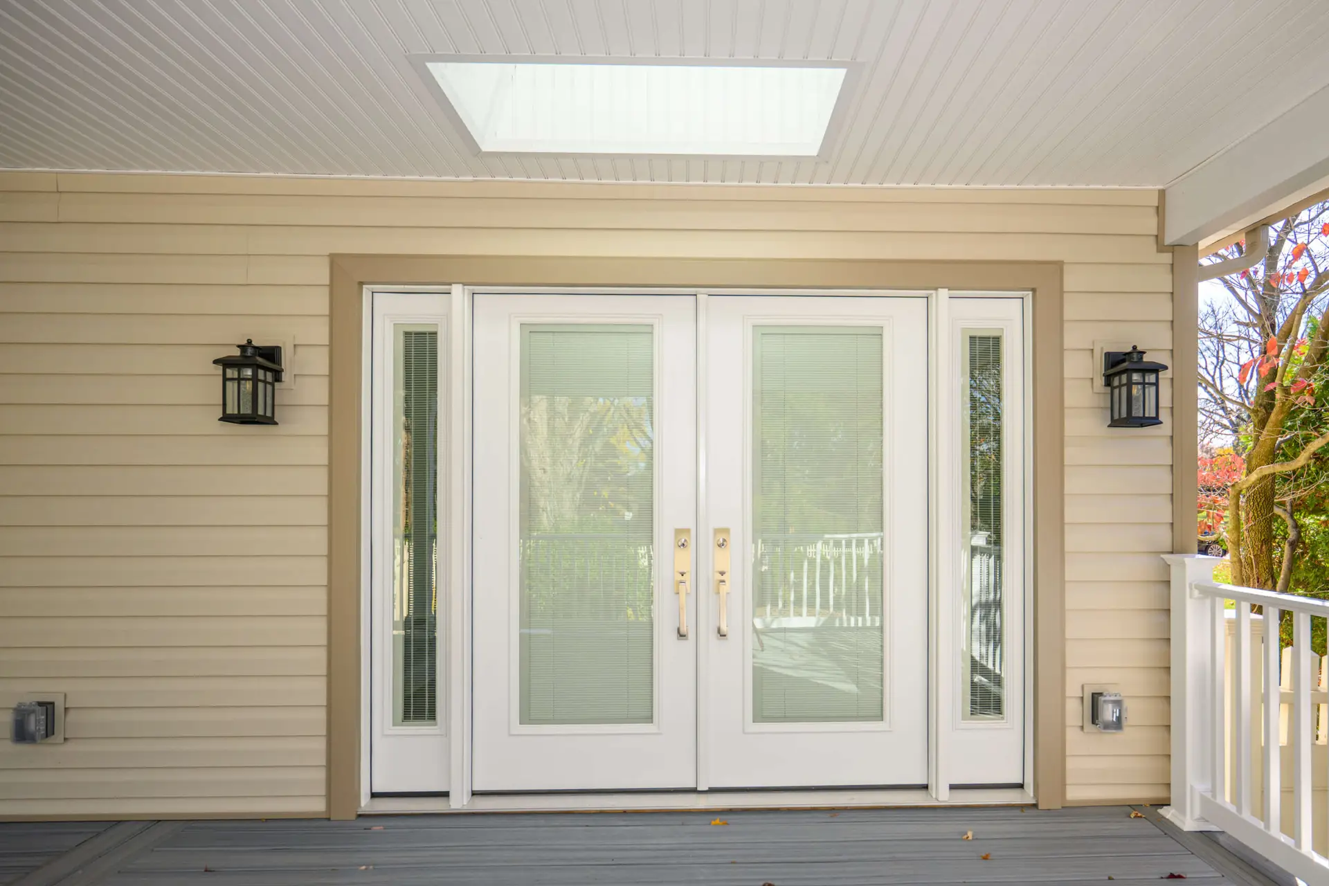 White double front doors with sidelights under a covered porch, flanked by black lantern sconces on beige siding. A skylight is above, with a gray wooden deck in the foreground.