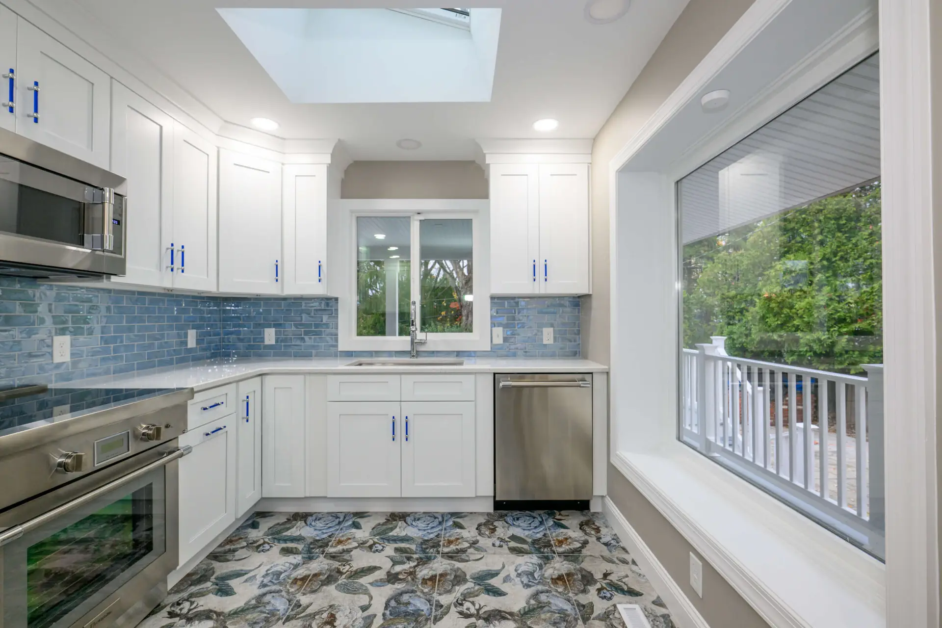 Bright, modern kitchen with white cabinetry, blue tile backsplash, and stainless steel appliances under a skylight and large window.