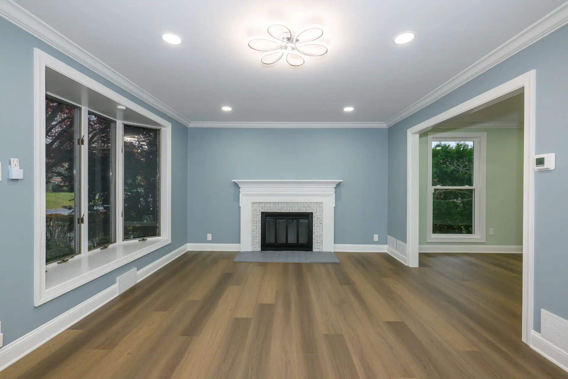 Bright living room with blue walls, white crown molding, a white fireplace with tiled surround, and wide windows on the left; wood plank flooring throughout.