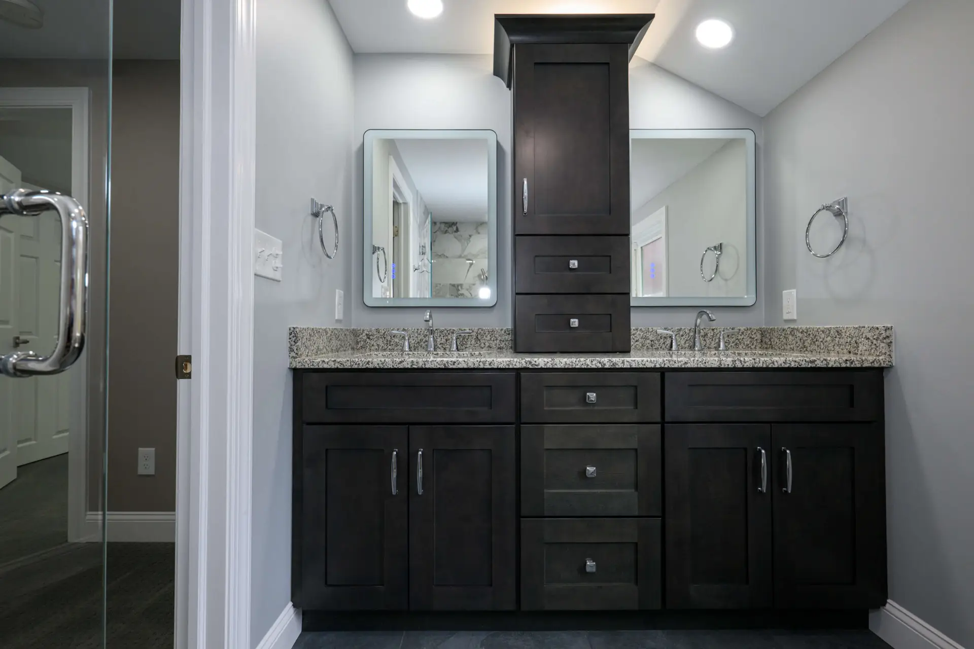 Dark wood, double-sink bathroom vanity with a speckled granite countertop, central tall storage tower, and two mirrors.