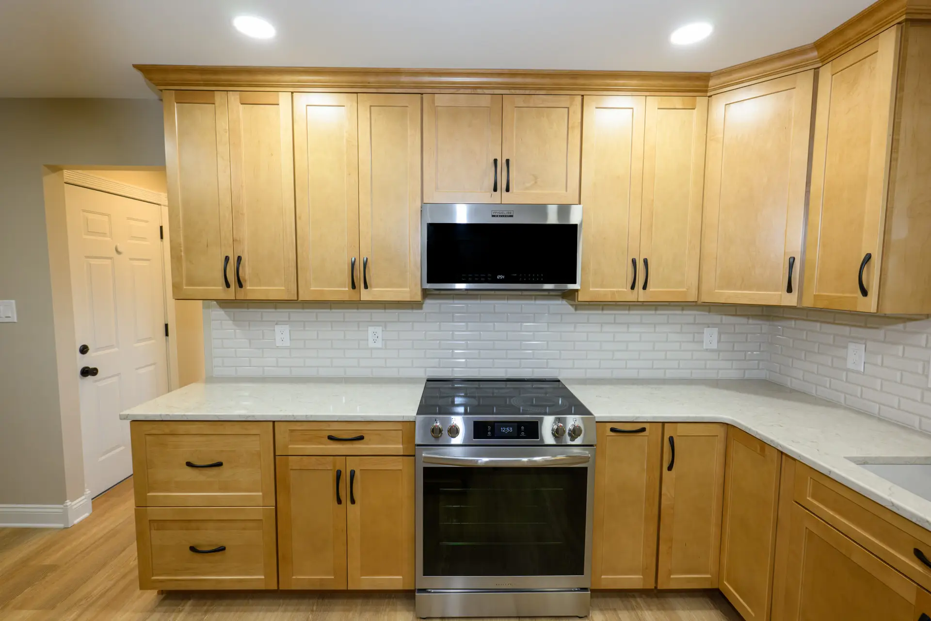 Kitchen with light wood cabinets and stainless steel oven, microwave above, and white tile backsplash; warm hardwood floor.