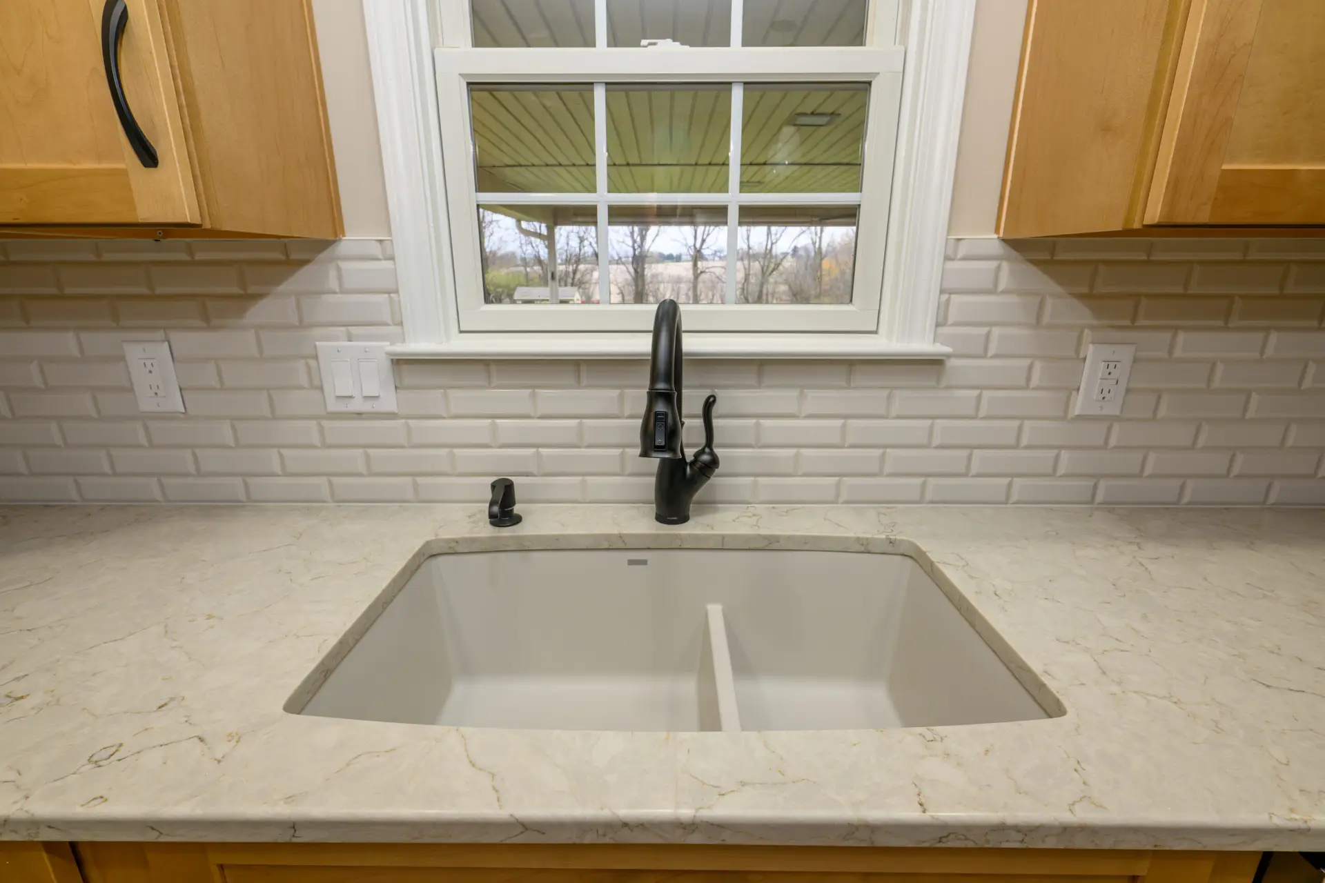 Black kitchen faucet over a white undermount sink with a marble countertop and white tile backsplash, window backdrop showing outdoor yard.
