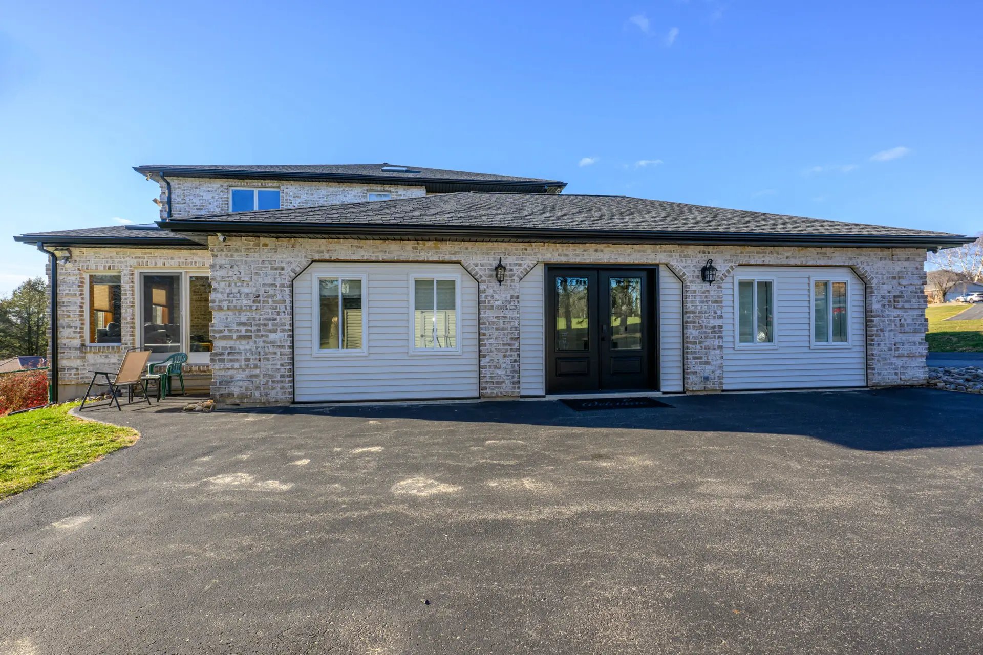 Front view of a brick house with a black double front door, white-trimmed windows, and a small seating area on the left under a covered porch, with a paved driveway in the foreground and blue sky above.
