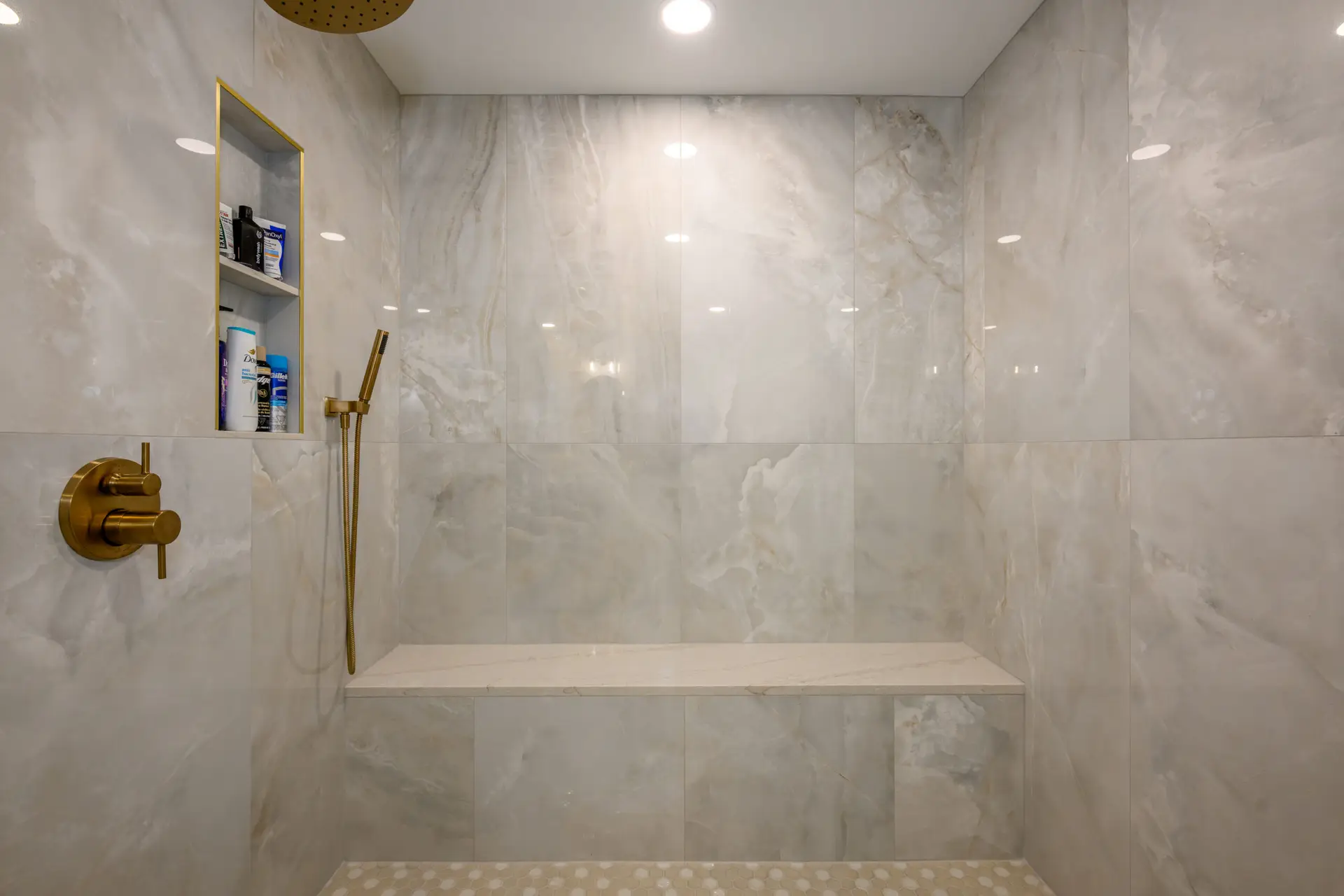 Elegant marble shower with a built-in niche stocked with toiletries, and gold bathroom fixtures with a wall-mounted showerhead and bench.