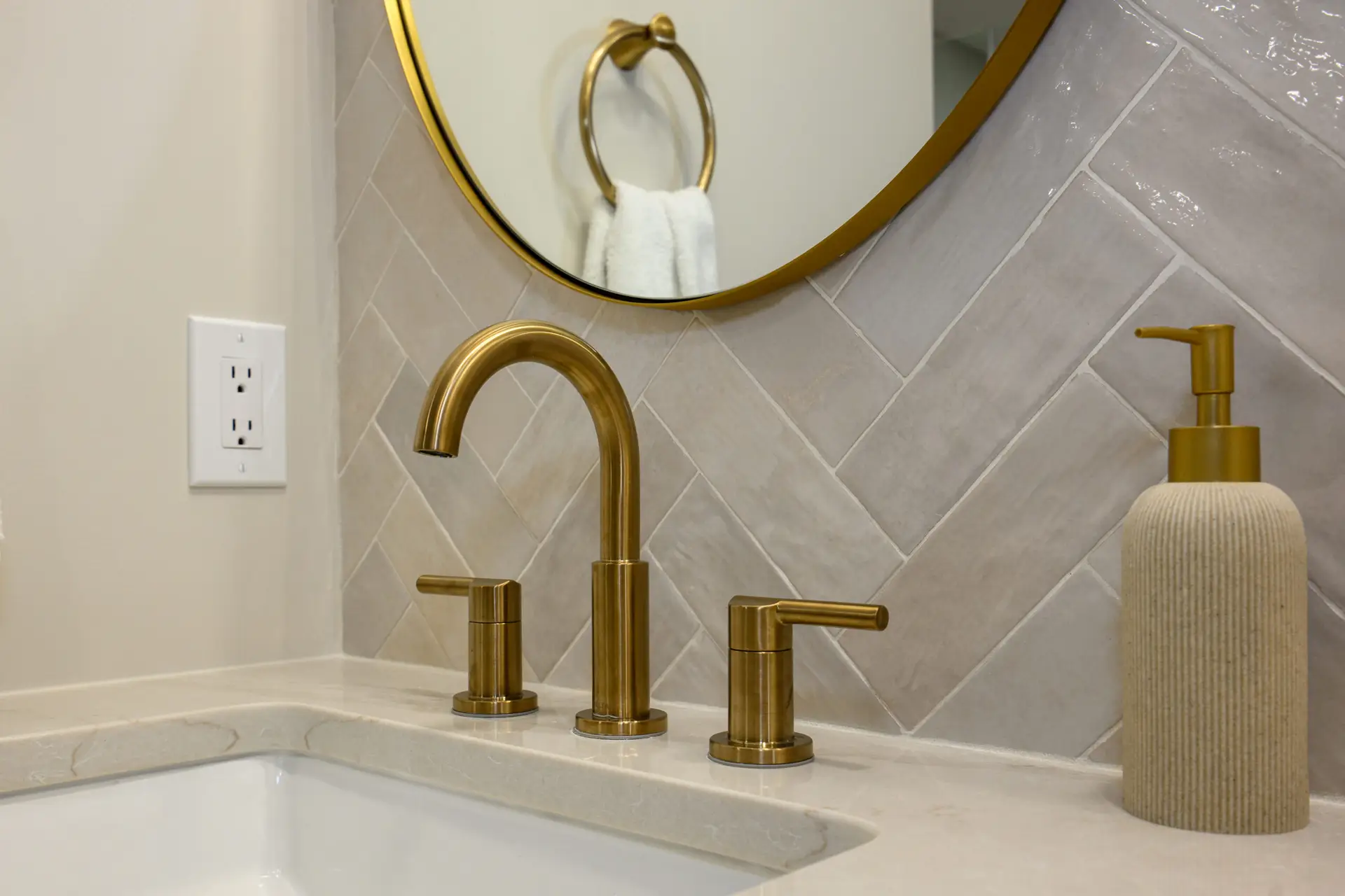 Bathroom vanity with two gold faucets, beige tiled backsplash, and a white towel on a gold ring near a round mirror.
