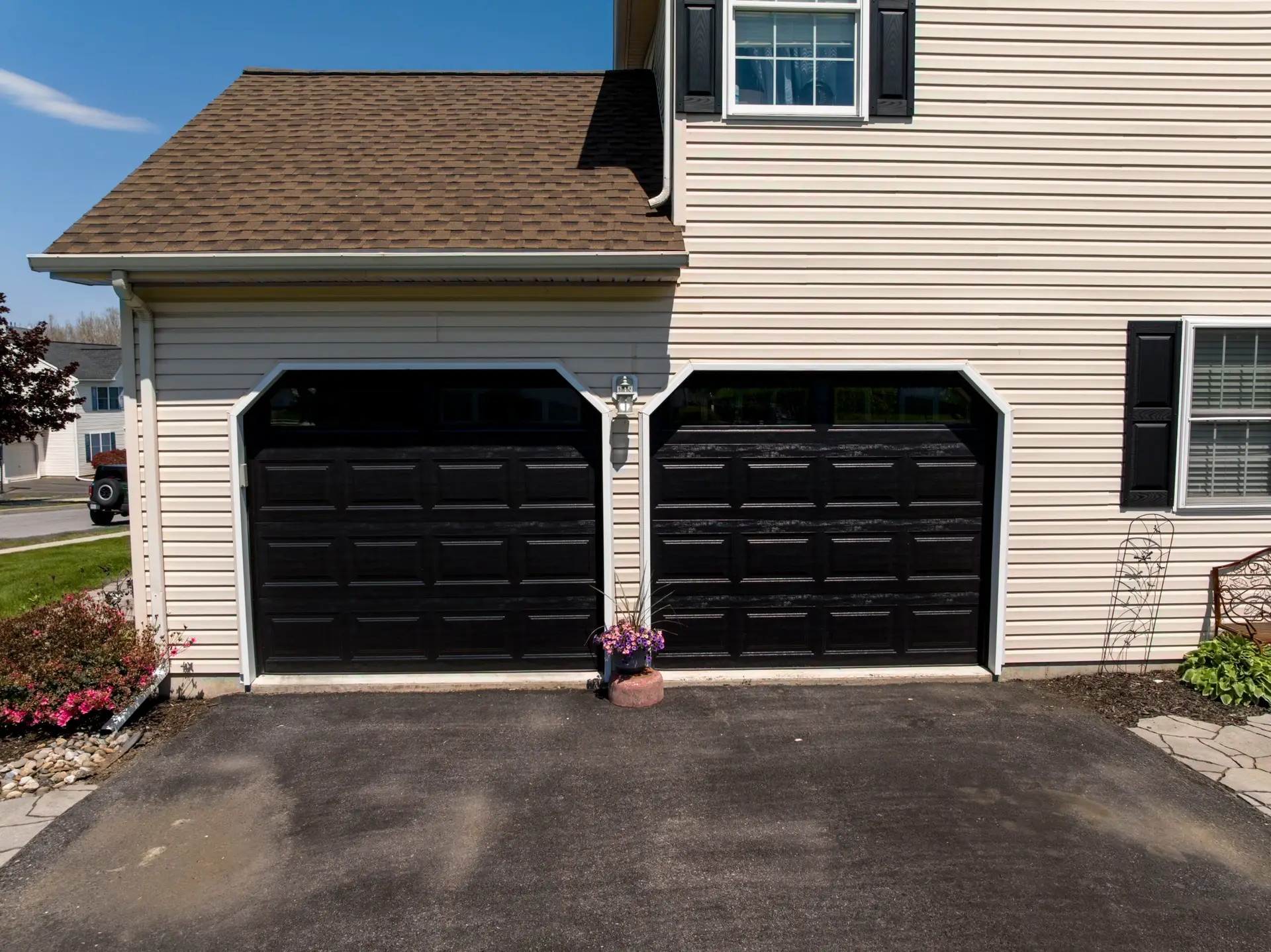 Front of a two-car garage with beige siding, brown shingle roof, and black doors; a flowering plant sits between doors.