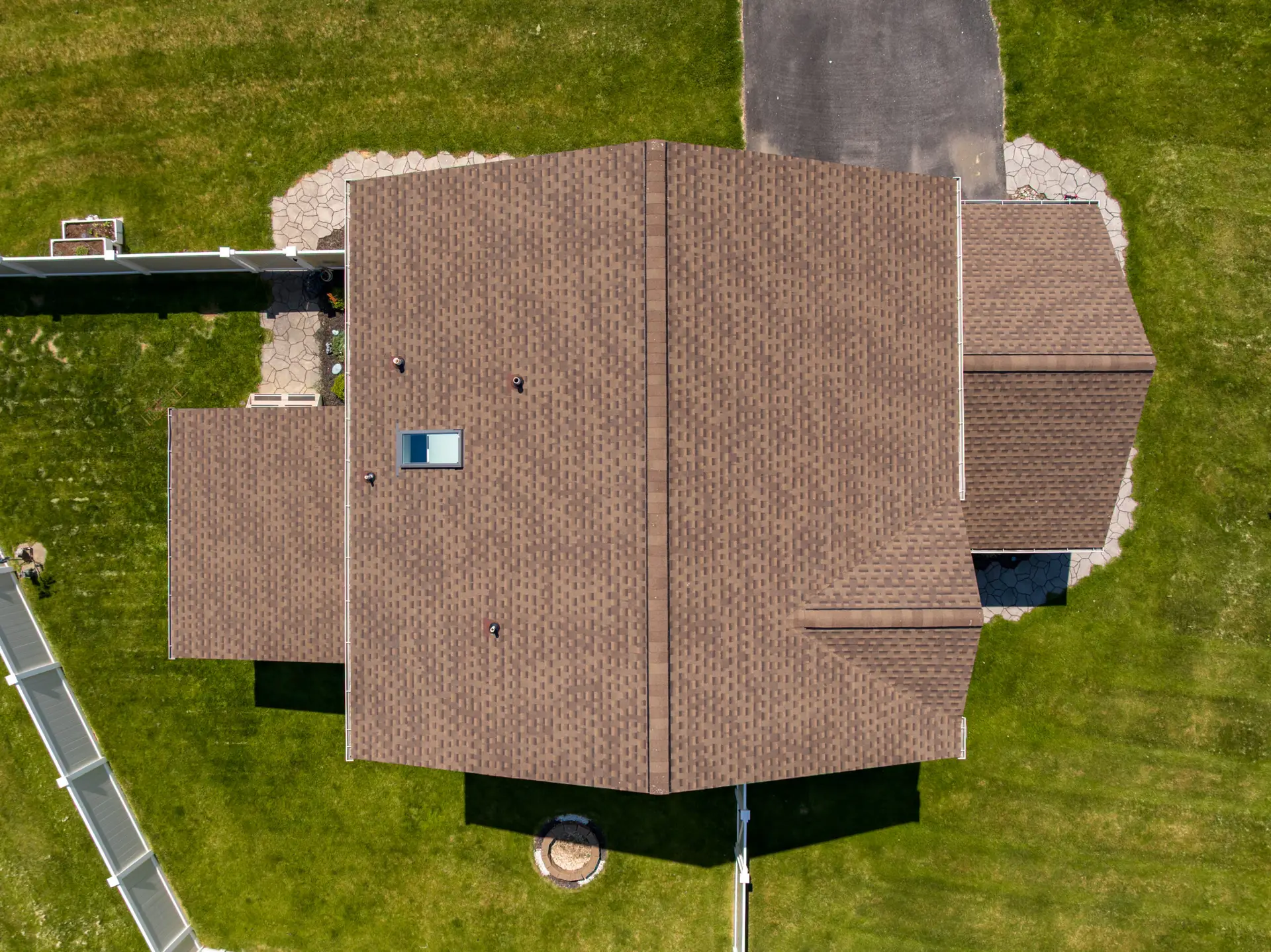 Aerial view of a large house with brown shingle roof, surrounded by green lawns and stone walkways. A circular stone feature sits in the lawn nearby.