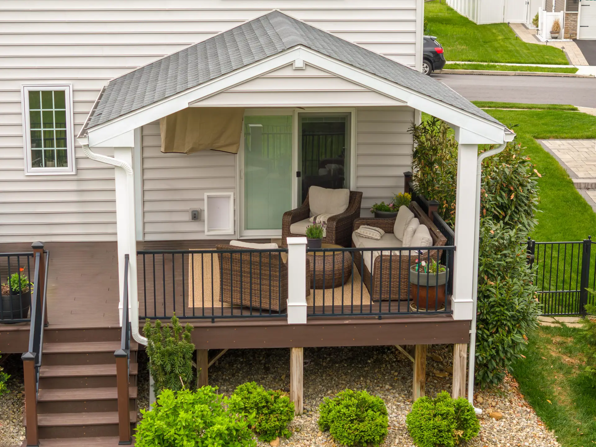 Front porch of a beige-siding house with a covered seating area, wicker sofa and chairs, and a glass sliding door on a wooden deck site.
