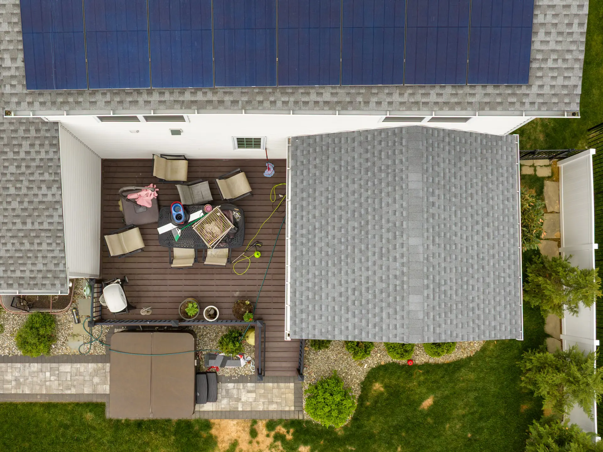 Top-down aerial view of a house roof with blue solar panels and a wooden deck featuring outdoor furniture.