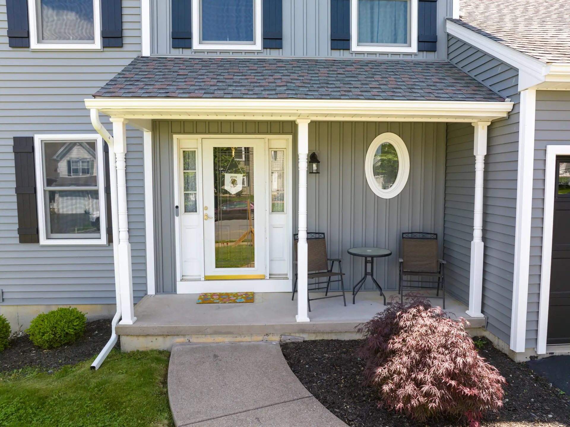 Front porch of a gray-blue house with white trim, glass double doors, a round window to the right, and outdoor seating on the porch.