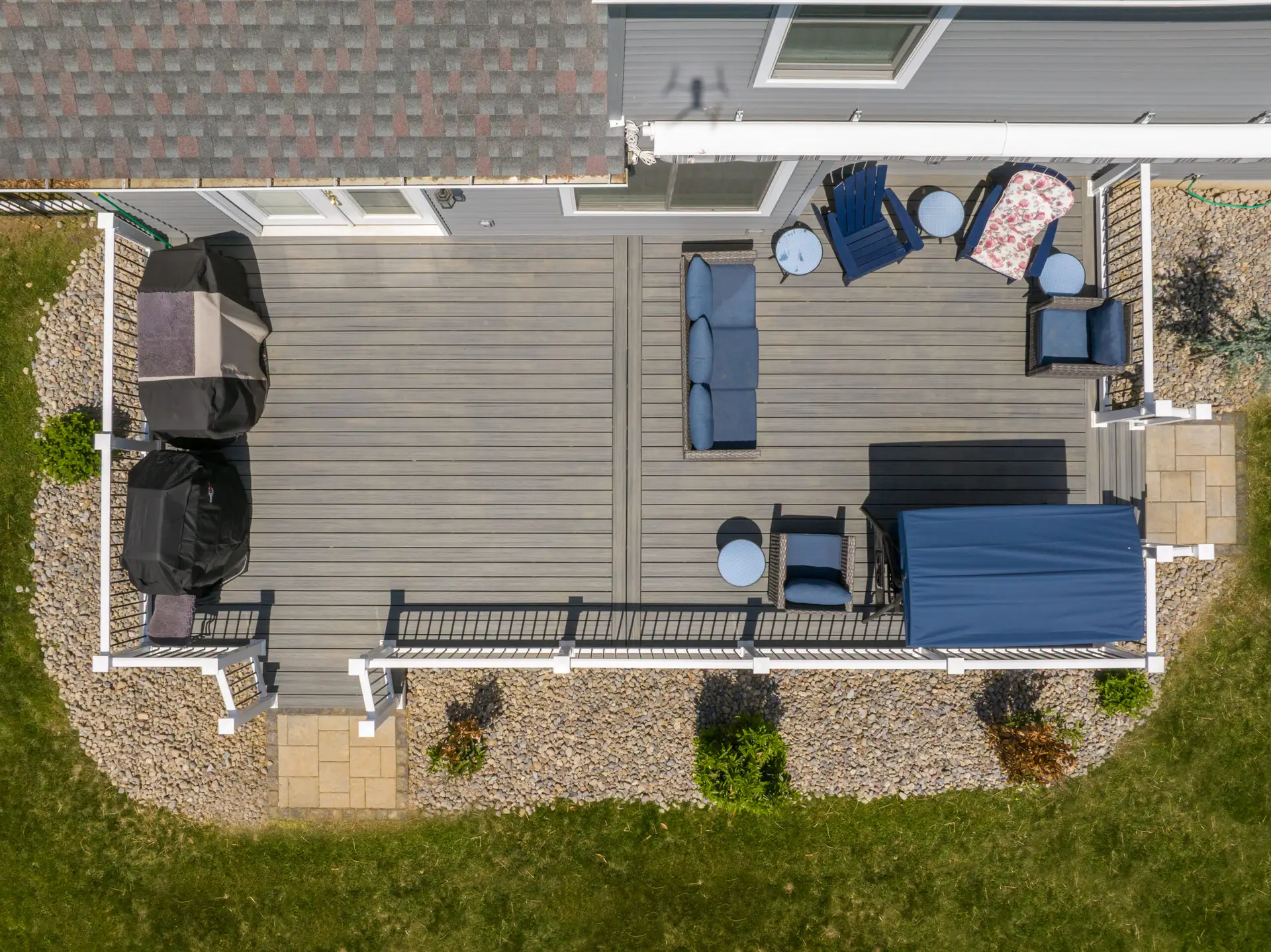 Aerial view of a wooden deck with blue patio furniture, a covered grill on the left, and a gravel border surrounding the yard.