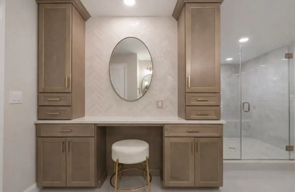 Beige bathroom vanity with tall cabinets, oval mirror, herringbone tile backsplash, and a cushioned stool at the center, with a glass shower to the right.
