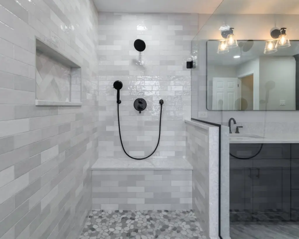 Glass-enclosed contemporary shower with gray subway tile walls, a large round rainfall showerhead and handheld wand, black fixtures, and a pebble-tile floor near a built-in bench.