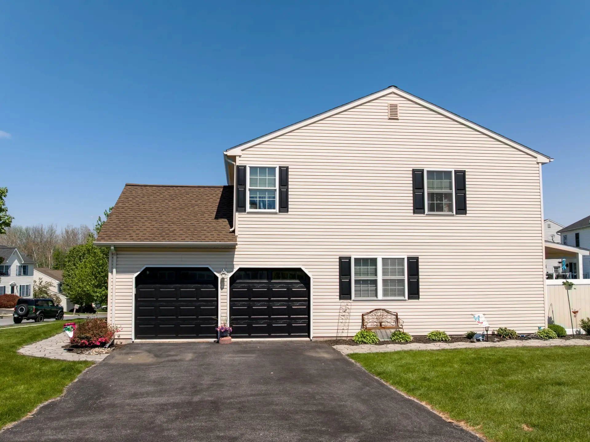 Two-story beige suburban house with black garage doors, white trim, and a curved driveway on a sunny day.