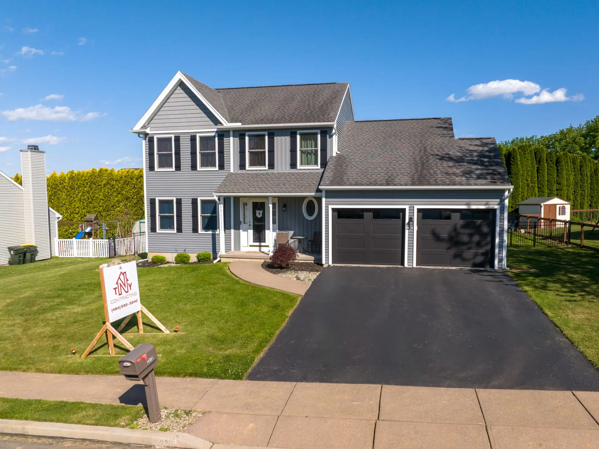 Front view of a gray two-story house with a dark, attached two-car garage and a manicured lawn; a contractor sign sits on the lawn in the foreground under a blue sky.