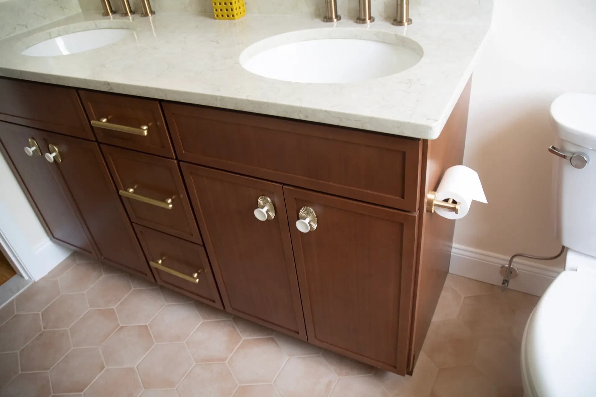 Double-sink bathroom vanity with a marble countertop, dark wood cabinetry, brass handles, and a toilet visible on the right.