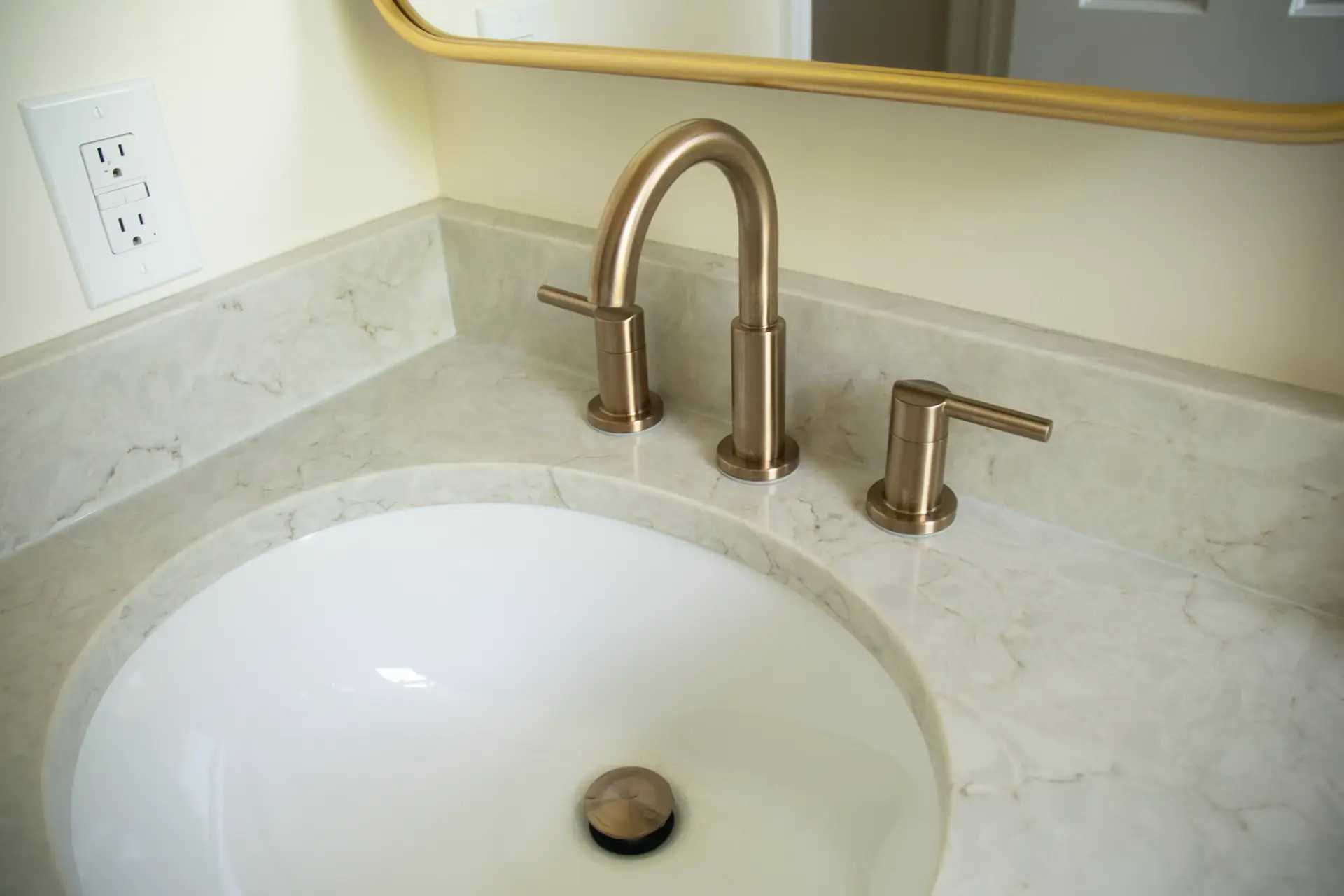 Bronze curved faucet over an oval white sink on a marble bathroom counter, with a wall outlet to the left.