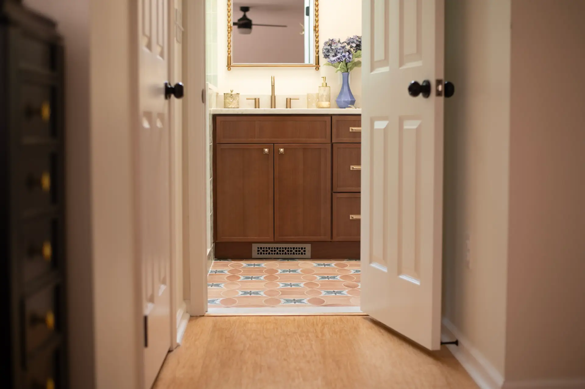 View into a bathroom with a wooden vanity, gold fixtures, and a blue vase of hydrangeas on the counter, seen through an open doorway.