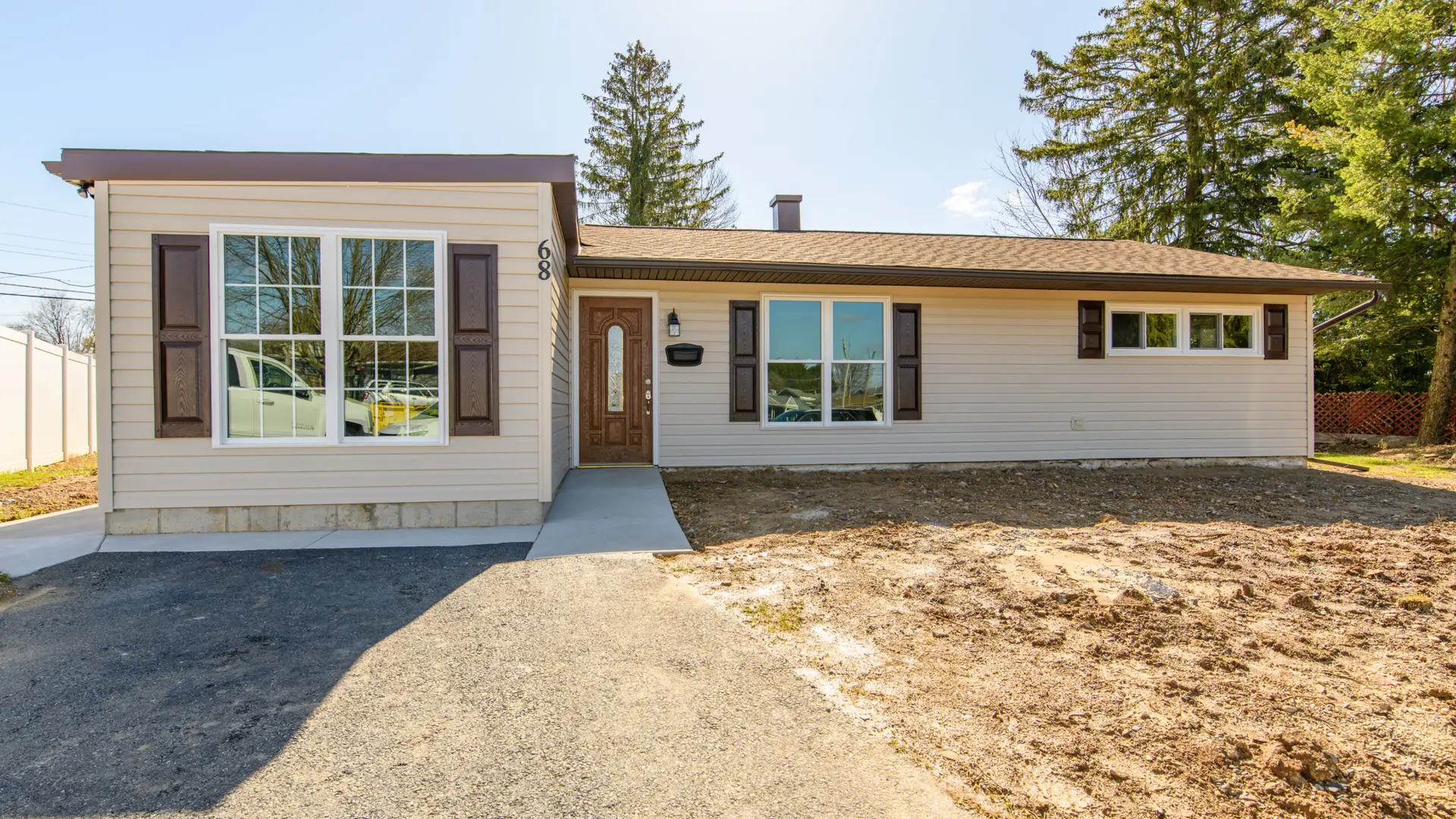 Front view of a beige single-story house with brown shutters, a wooden entry door, and a large window; dirt yard in front.