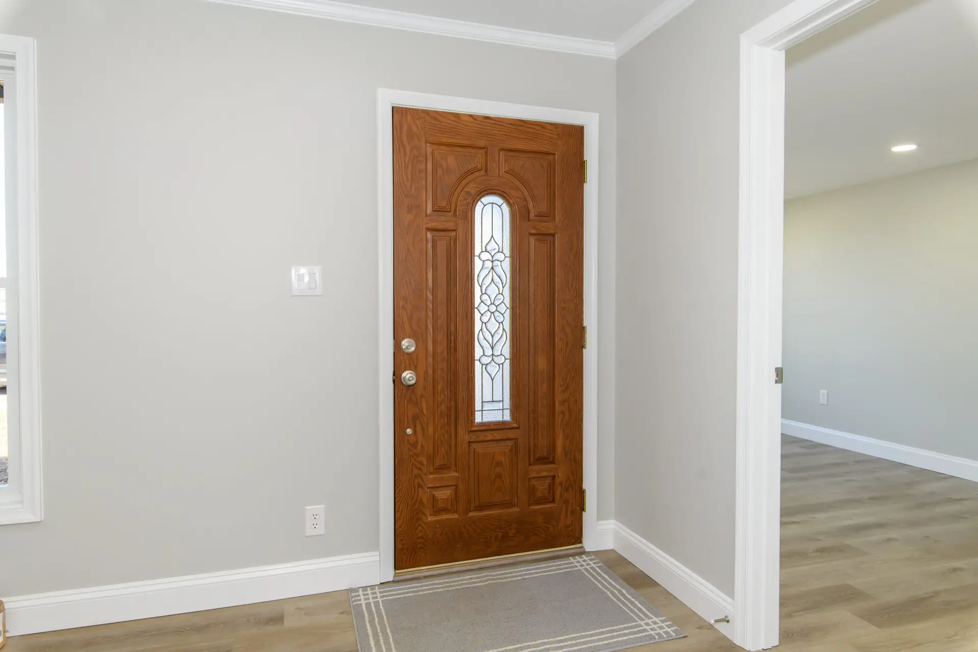 Wood front door with decorative glass panel in a light gray entry hall, beige rug on laminate floor.