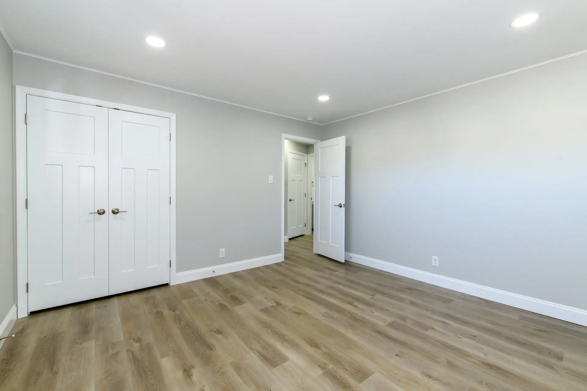 Empty modern room with gray walls, white double closet doors, and an open doorway; wood-look flooring and recessed ceiling lights visible.