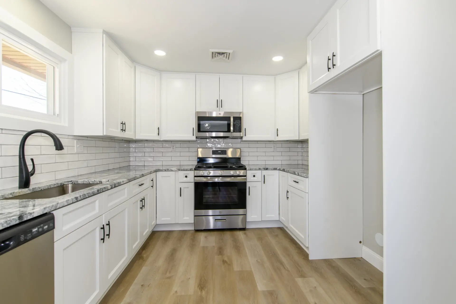 Modern white U-shaped kitchen with stainless steel oven, microwave, and granite countertops, plus a black faucet at the left sink.