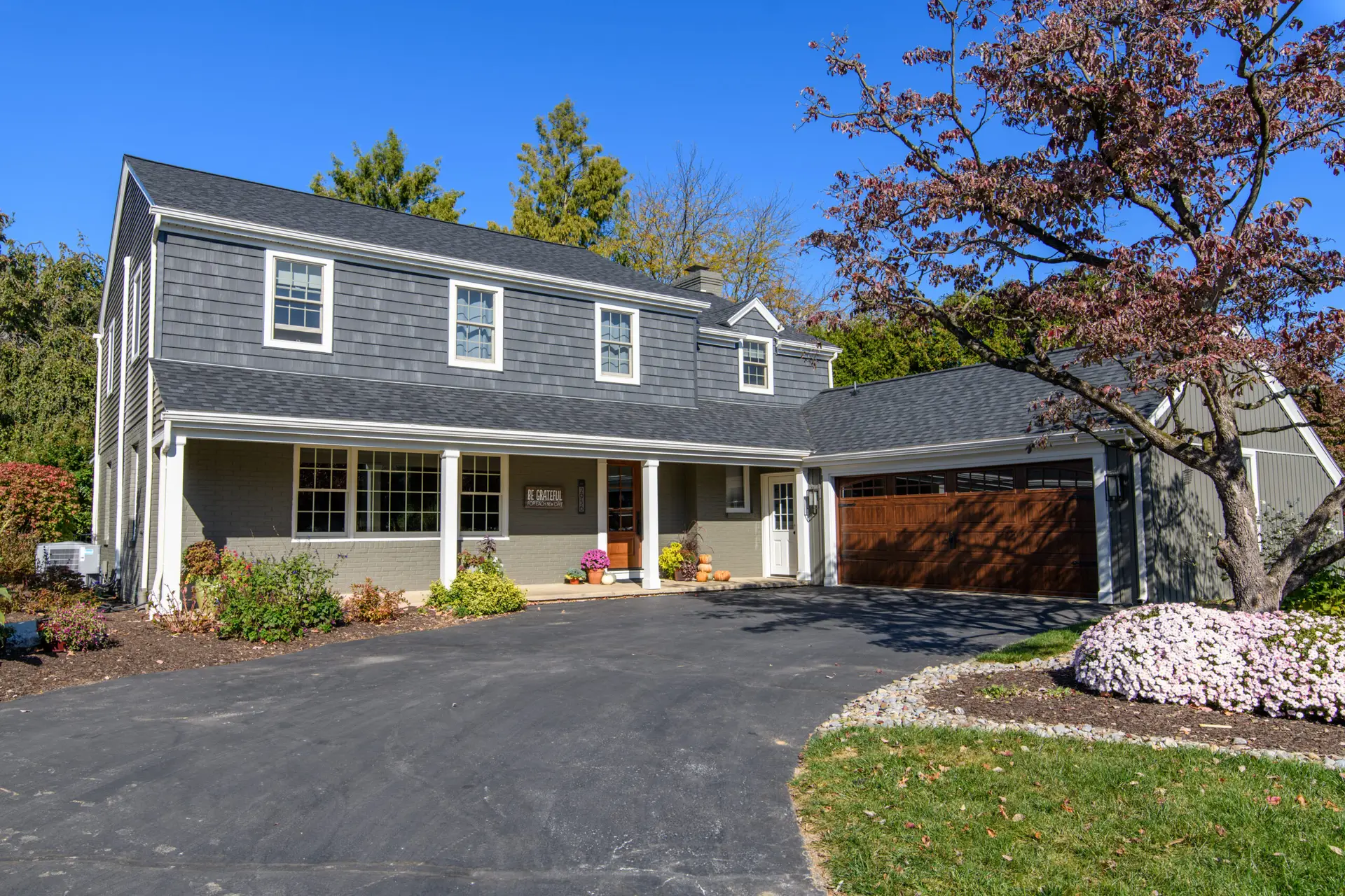 Front view of a gray two-story house with dormer windows, a brown garage door, and autumn decorations on the porch under a bright blue sky.