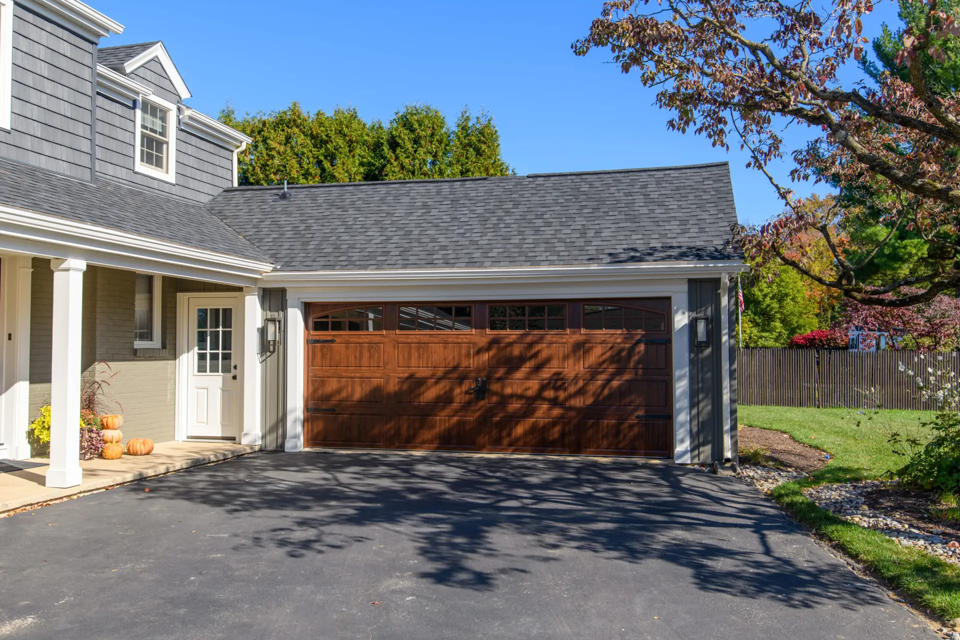 Front view of a suburban home with a gray shingle roof, white trim, and a wooden garage door; pumpkins sit on the porch in autumn light.
