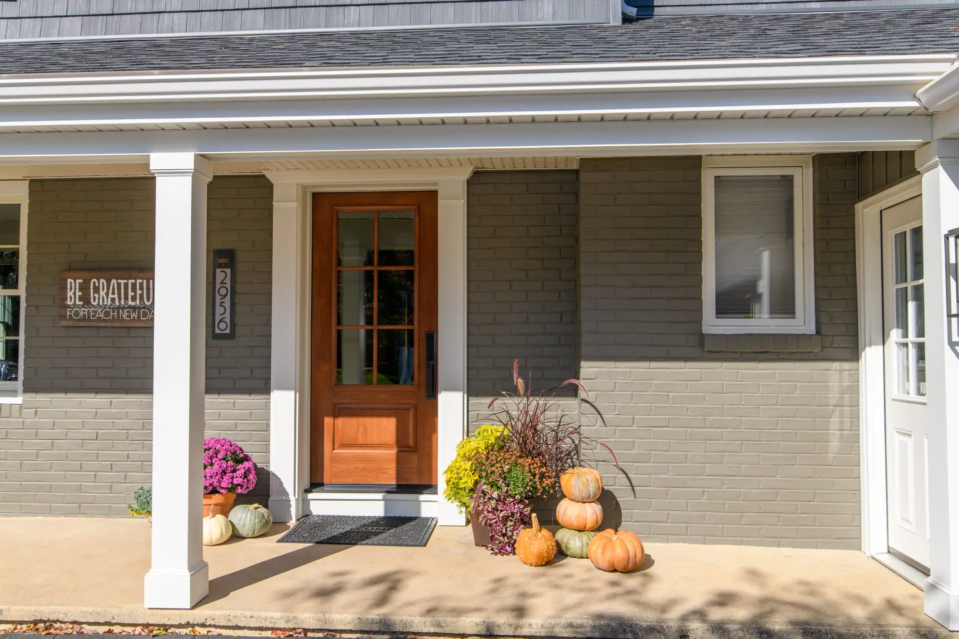 Front porch of a gray brick house with white columns, a wooden front door, and fall decorations: pumpkins and mums scattered around the entryway.