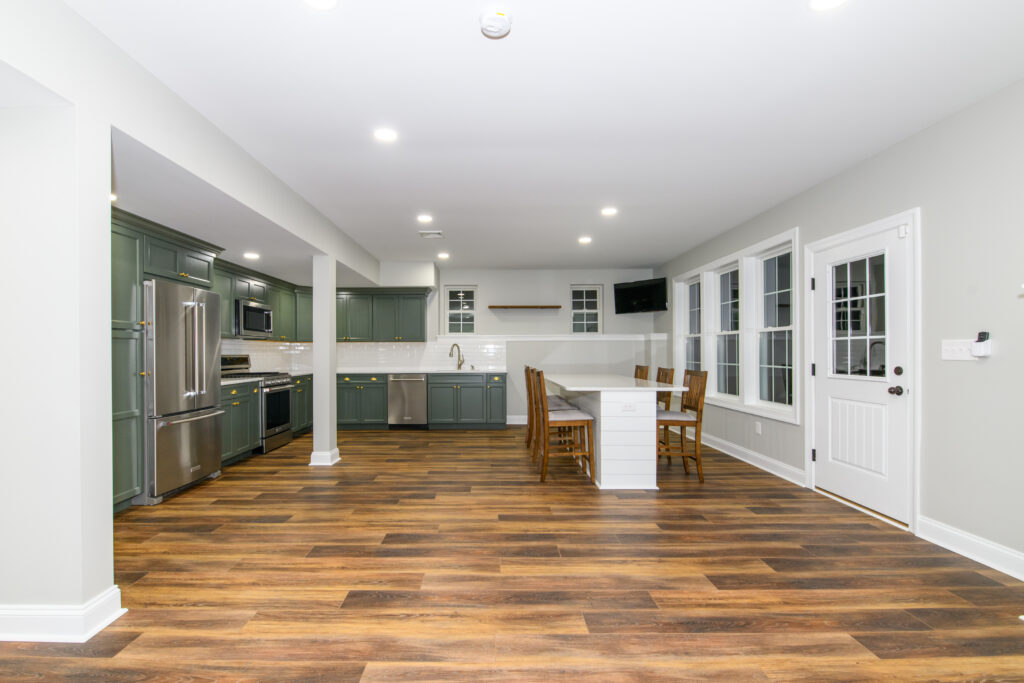 Open-concept kitchen with green cabinets, stainless appliances, and a white island with seating on a hardwood floor; dining chairs nearby beyond.