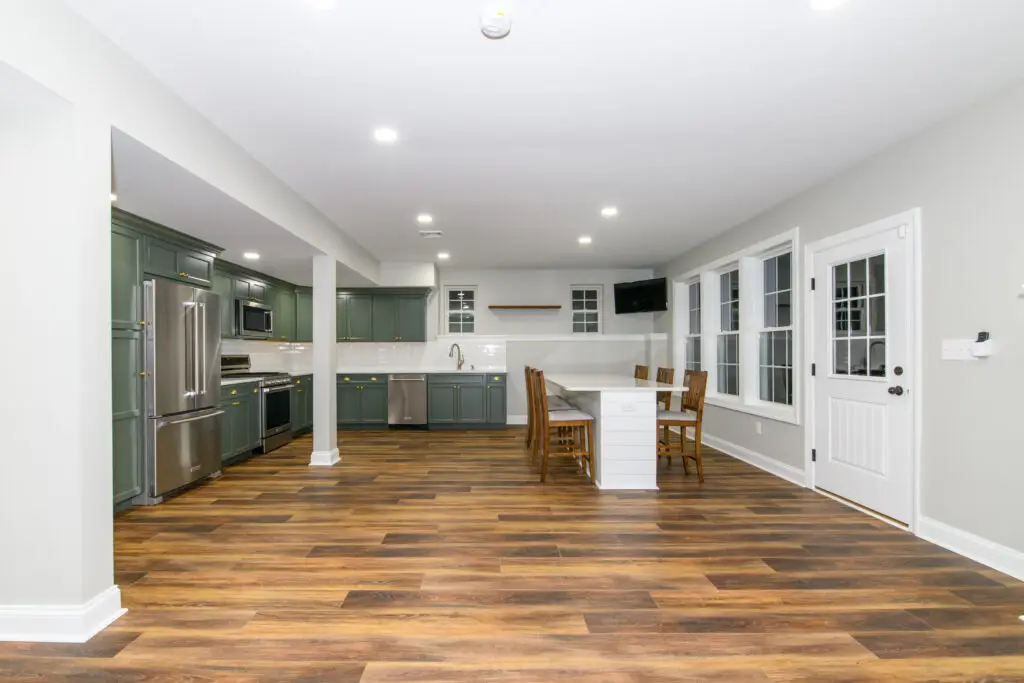 Open-concept kitchen with green cabinets, stainless appliances, and a white island with seating on a hardwood floor; dining chairs nearby beyond.