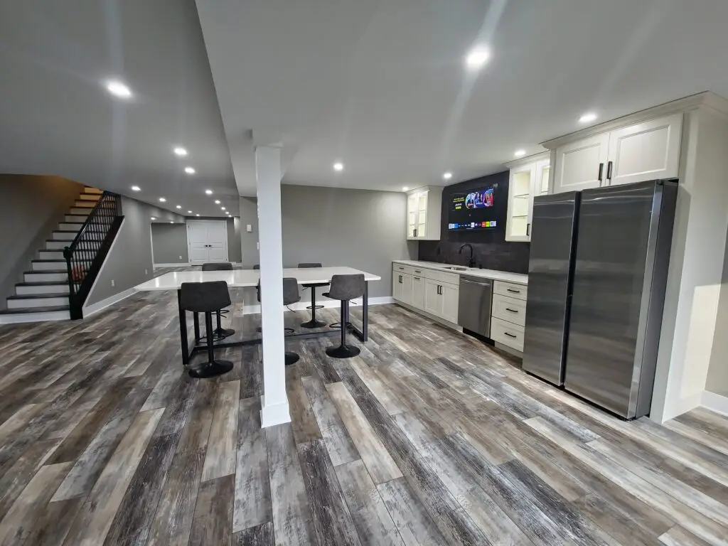 Open-concept kitchen with white cabinets, stainless steel French-door fridge, and a central island with bar-height chairs; stairs visible on the left.