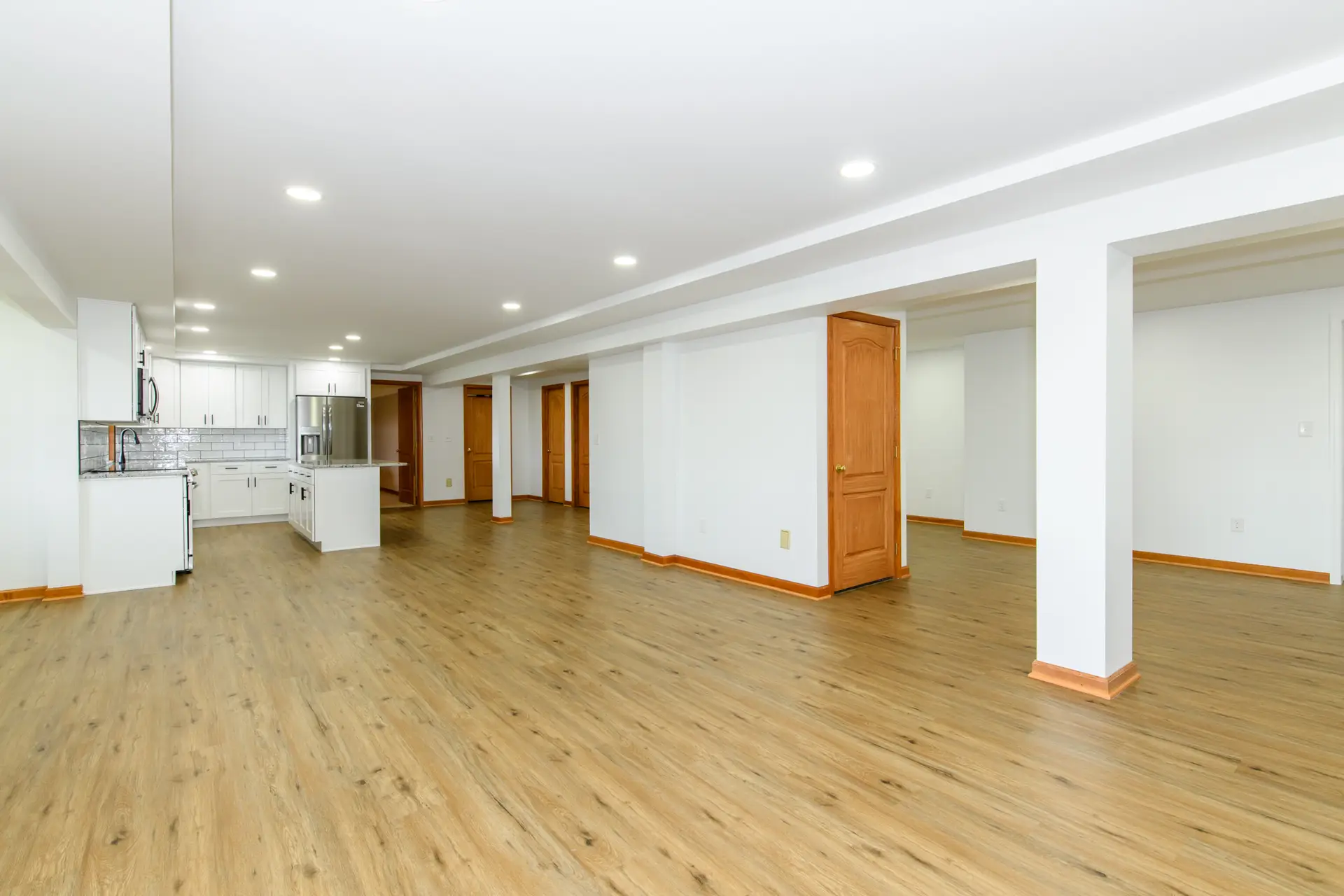Open-concept basement with a white kitchen on the left, warm wood floors, and multiple wooden doors along a white wall.