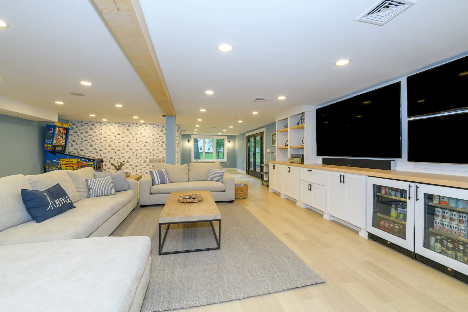 luxury-finished-basement-lehigh-valley - TNY Contracting - TNY Contracting Bright basement living room with a beige sectional, wooden coffee table, light-blue walls, and white built-in cabinets with a beverage fridge and large black TVs.