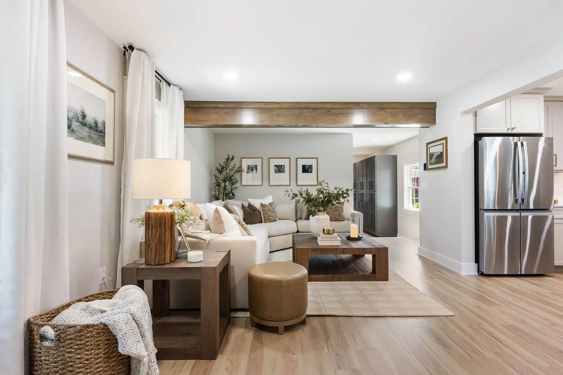 Bright open-concept living room with beige sectional, wooden coffee table, and neutral decor; French doors with white curtains frame the space.