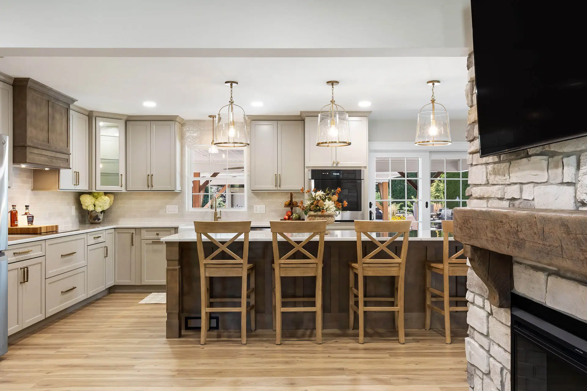 Bright kitchen with white cabinets, a central island, and three pendant lights; wooden bar stools aloft, stone fireplace on the right edge.