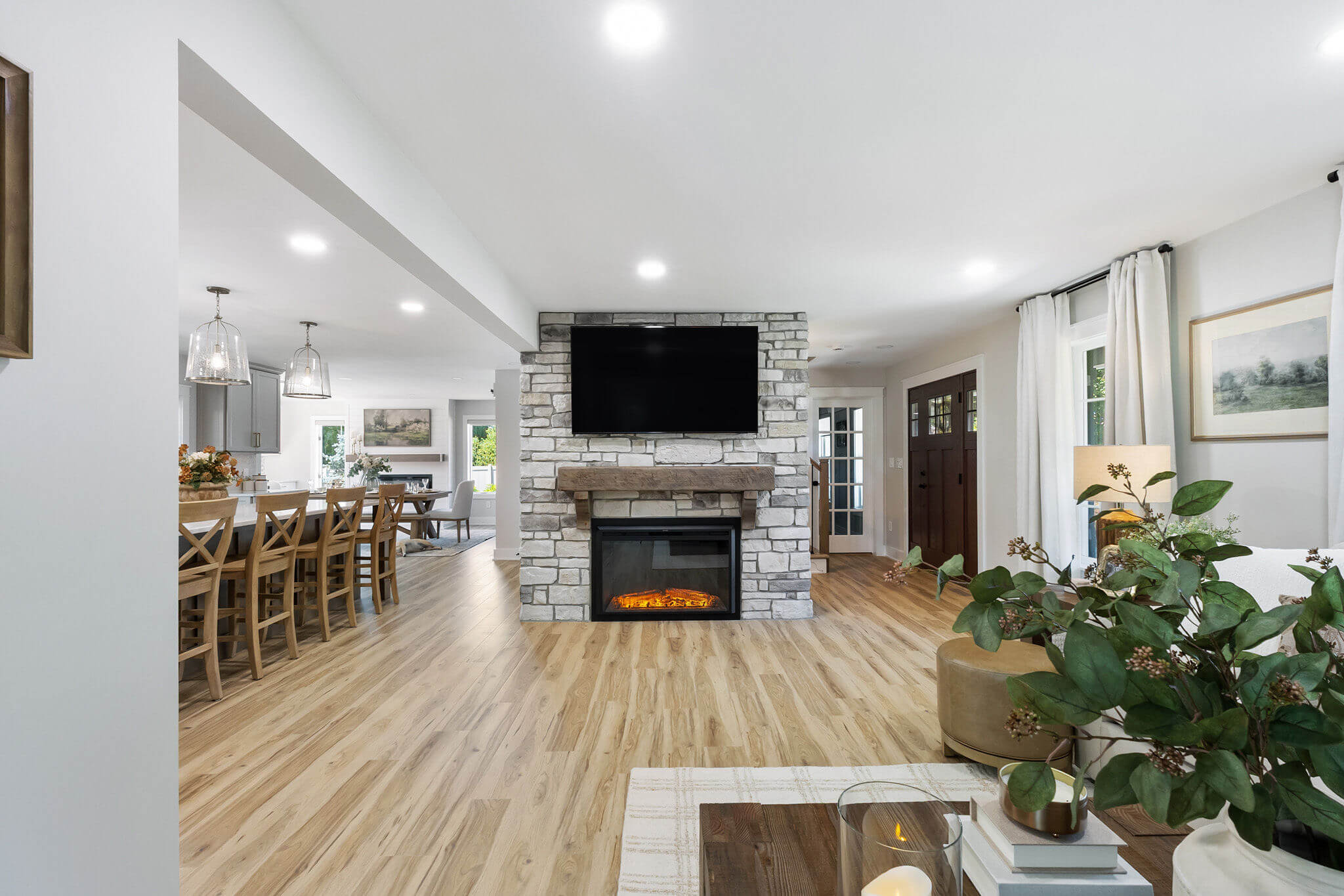 Bright open living room with a stone fireplace, mounted flat-screen TV, and dining area to the left in a light-filled space.