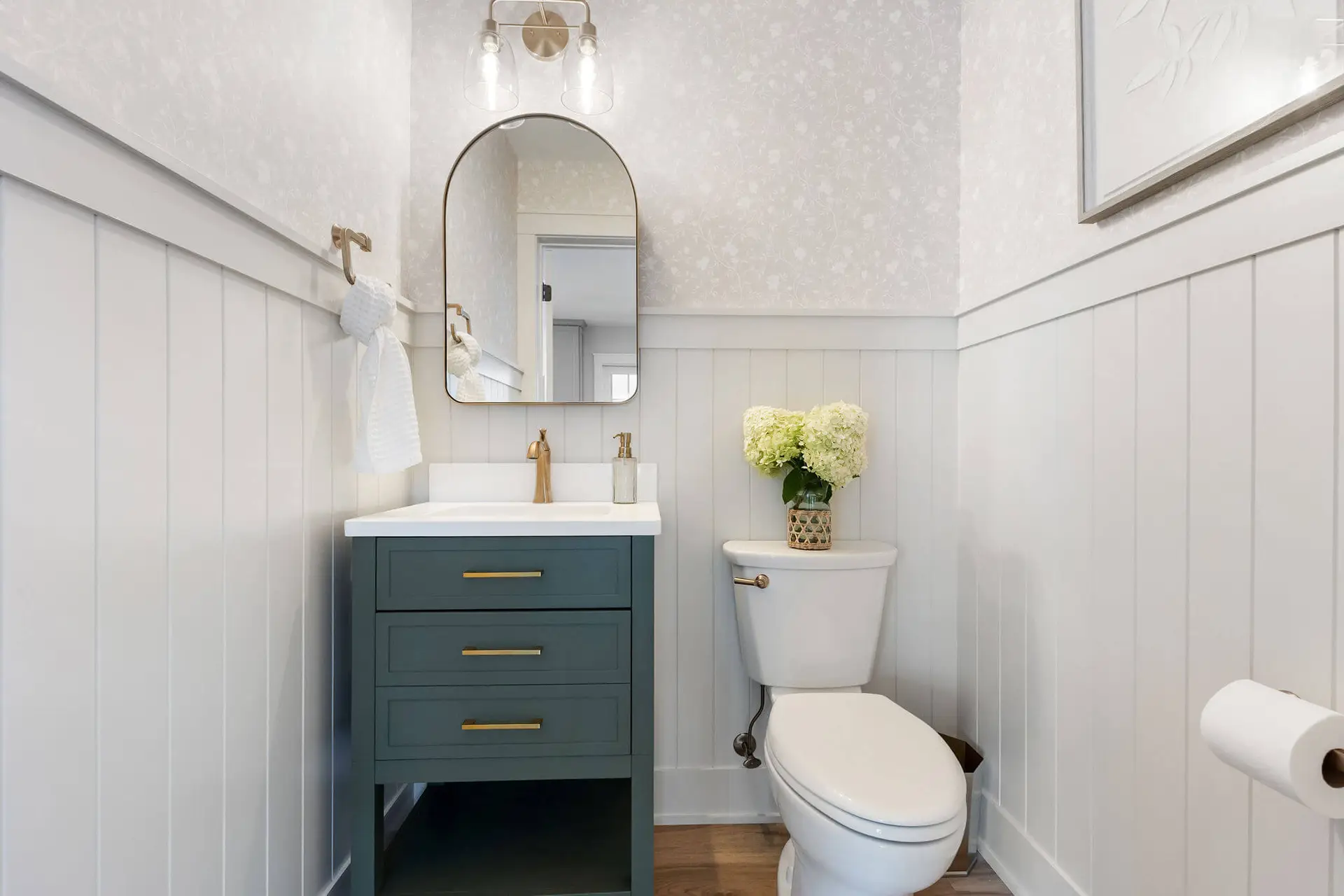 Compact bathroom with white beadboard wainscoting, teal vanity, gold faucet, round mirror, and a vase of white hydrangeas on the toilet tank.