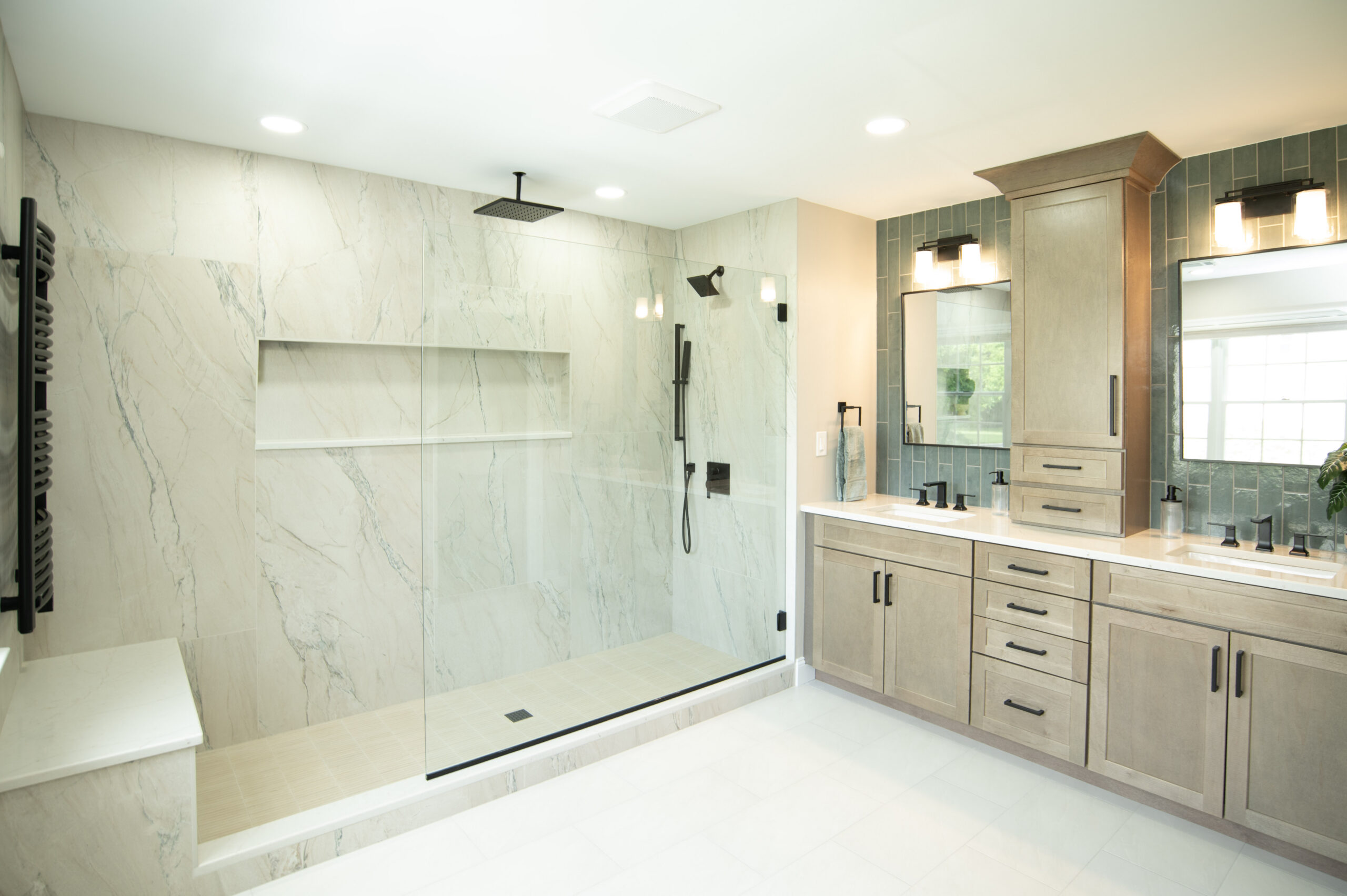 Glass-walled walk-in shower with marble walls and rainfall ceiling showerhead beside a double vanity with wood cabinets and black fixtures.