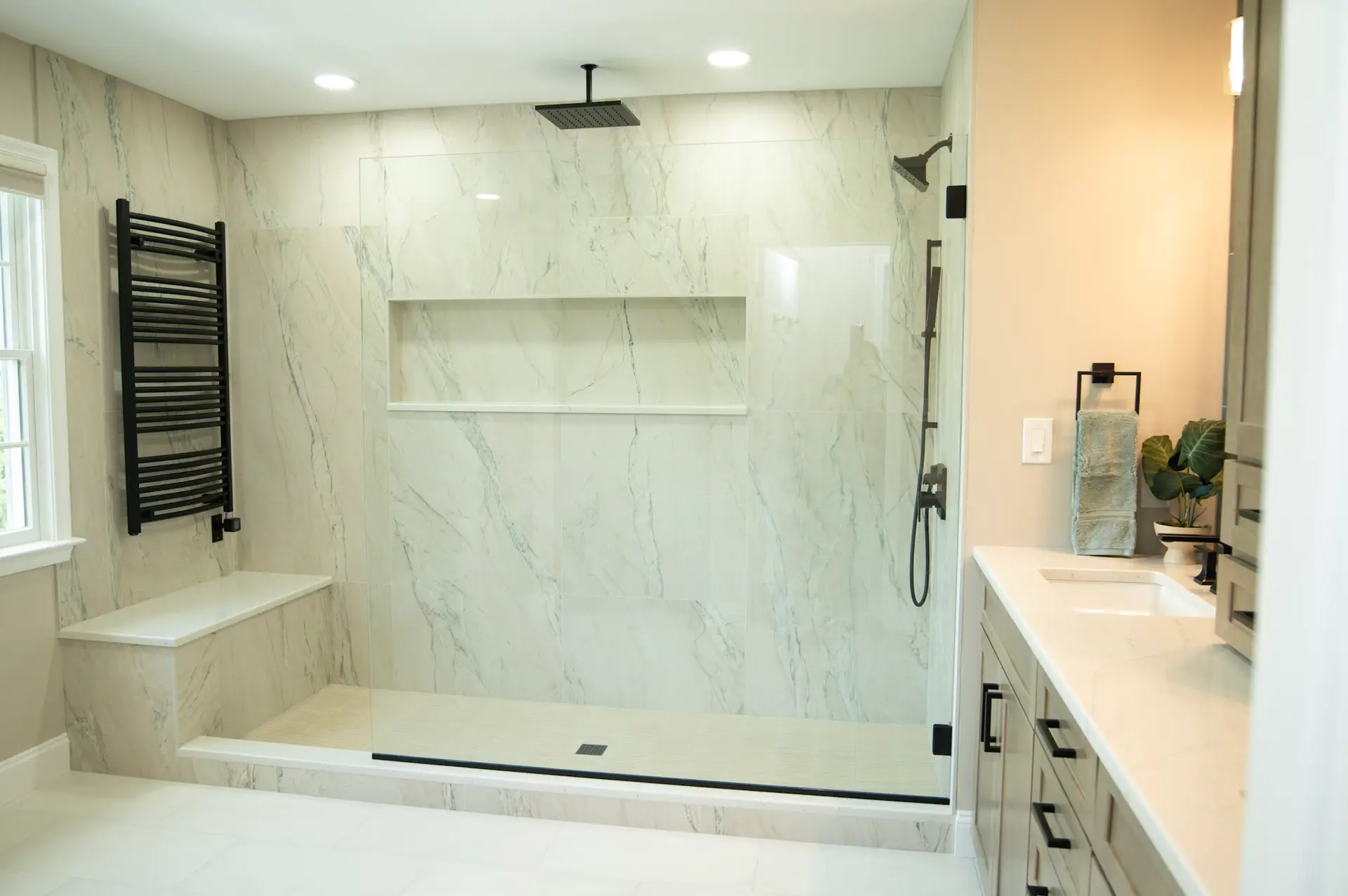 Spacious marble-tiled bathroom with a glass walk-in shower, black heated towel rack, and a long vanity on the right.